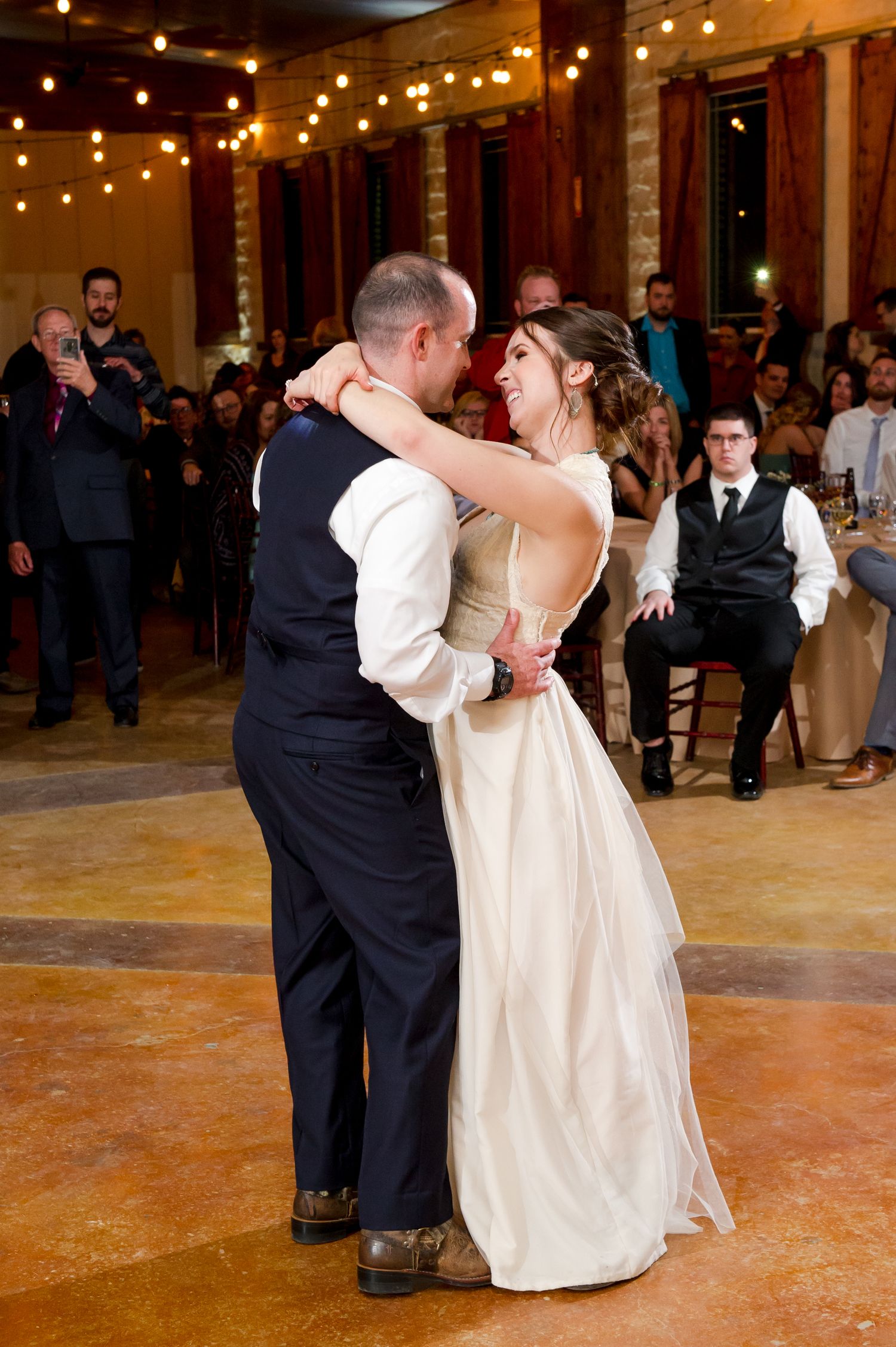 Couples dance together at an indoor wedding reception under warm string lights on a rustic wooden floor.