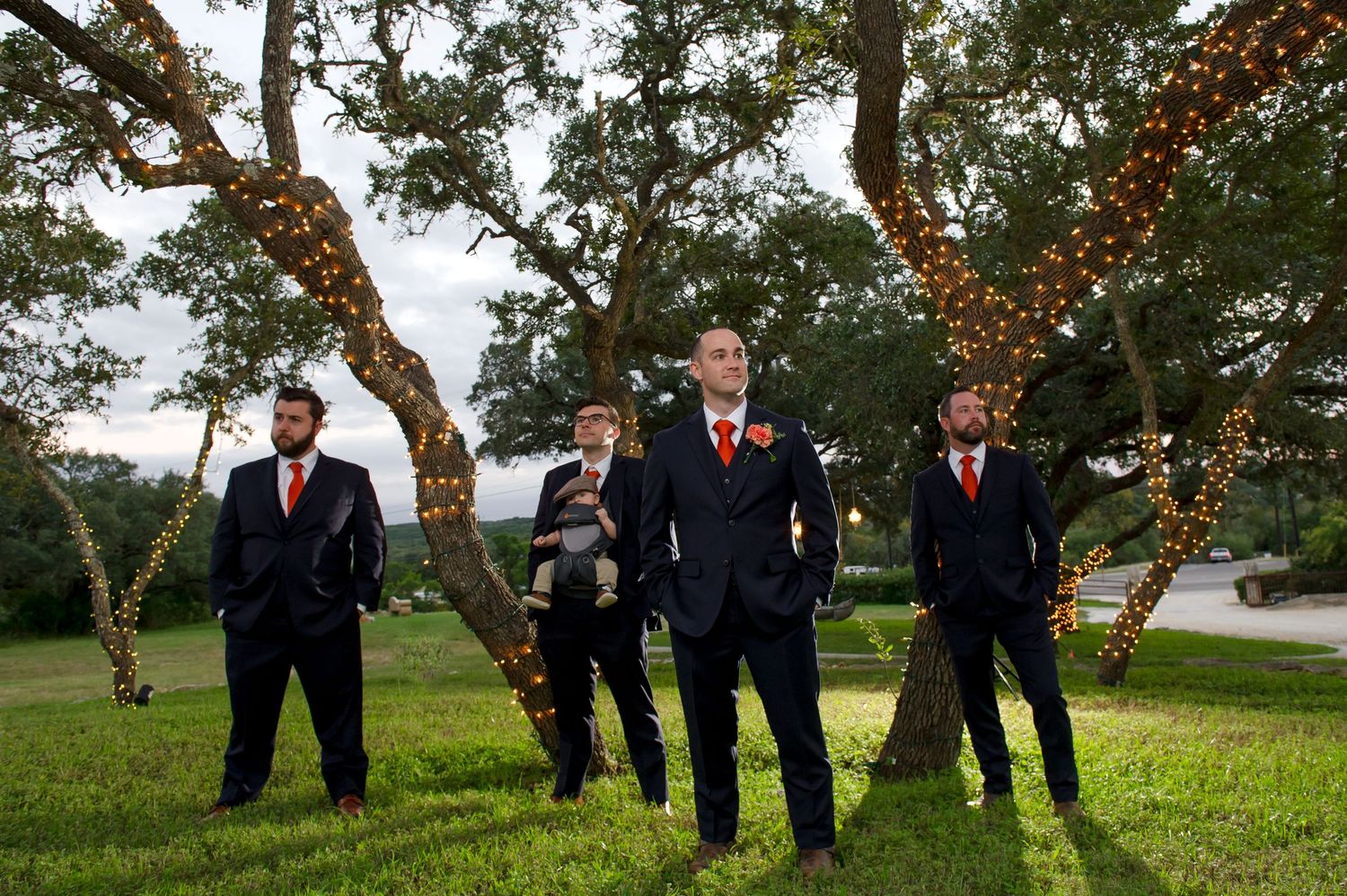 Wedding party poses under trees decorated with hanging lights in outdoor setting.