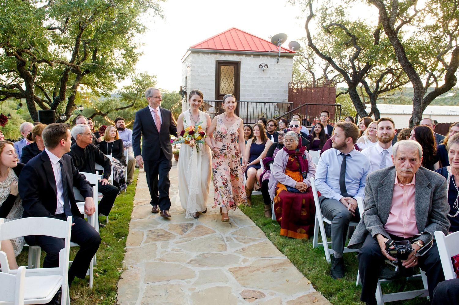 Wedding ceremony guests seated outdoors at a garden venue with a small gazebo and stone pathway.