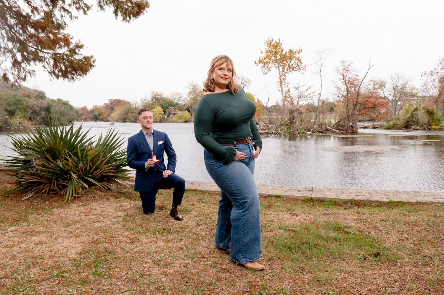 A couple poses for engagement photos near a lake with fall colors in the background.