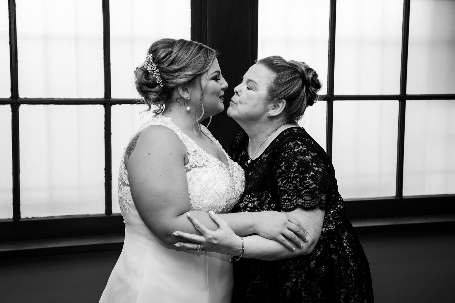 Two people in formal attire share an intimate moment in front of a window with black and white lighting.