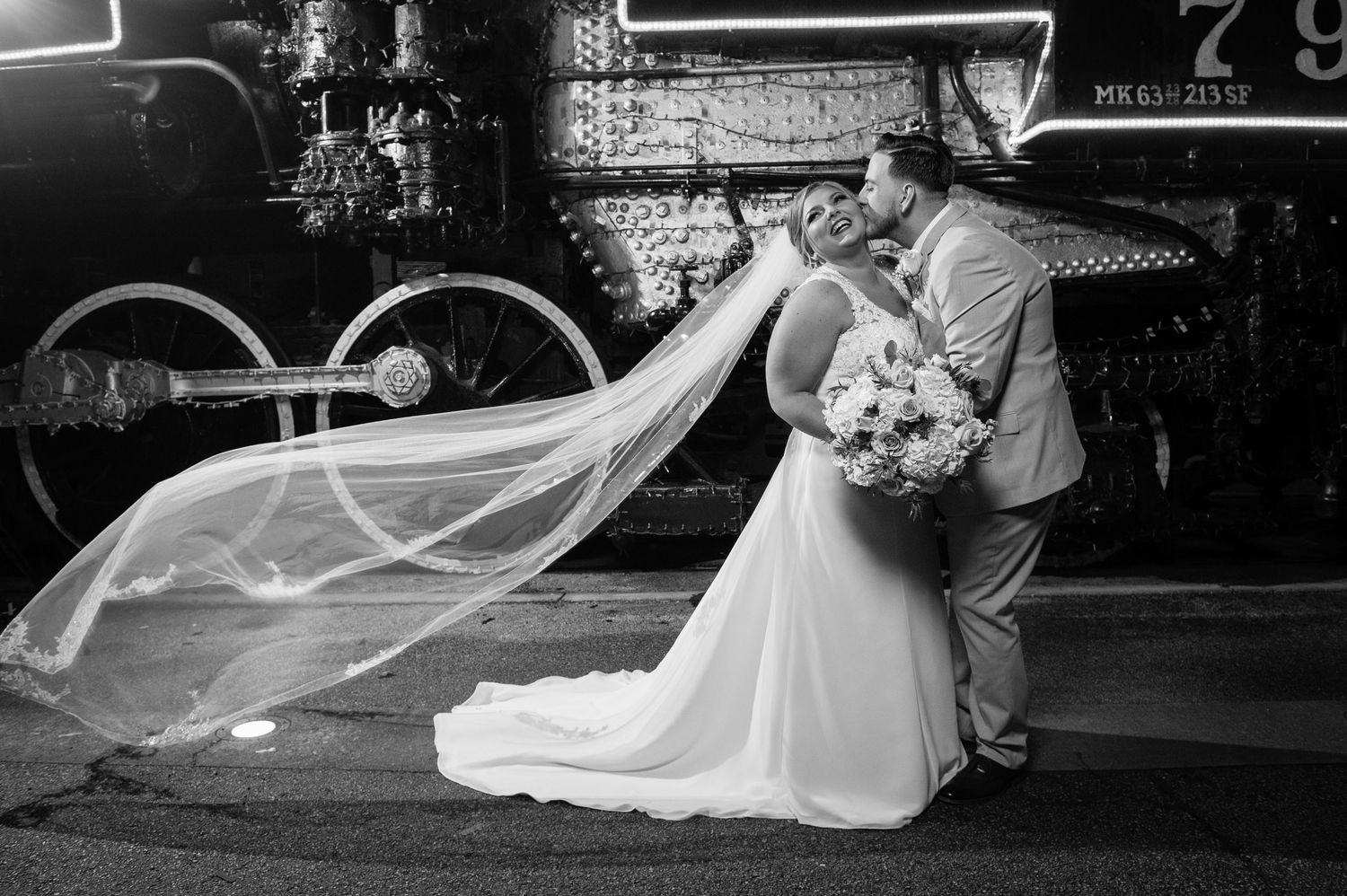 Elegant wedding veil flows in the breeze against industrial railway backdrop.