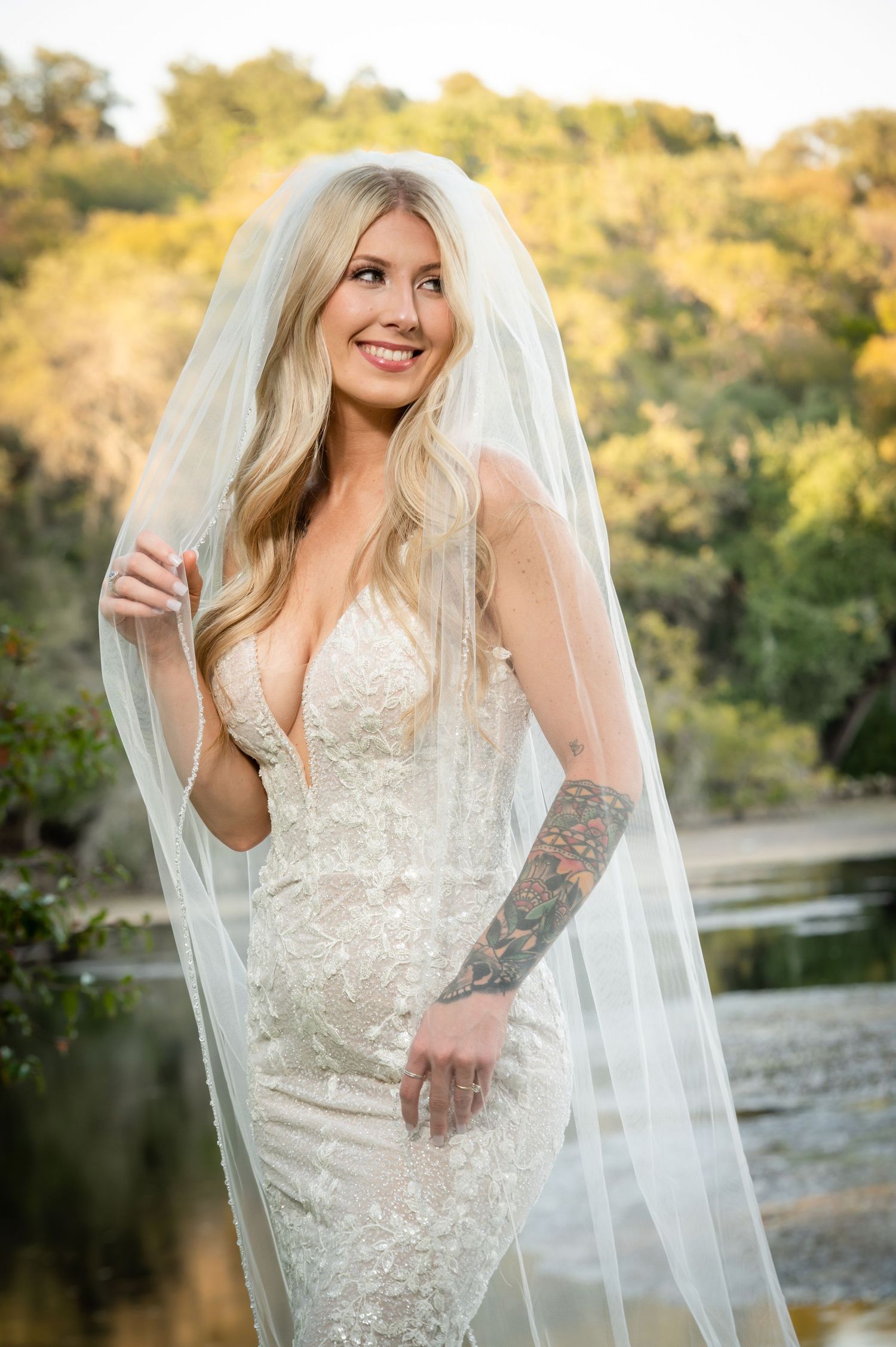 A bride in a detailed lace wedding dress poses outdoors in golden sunlight with a flowing veil.