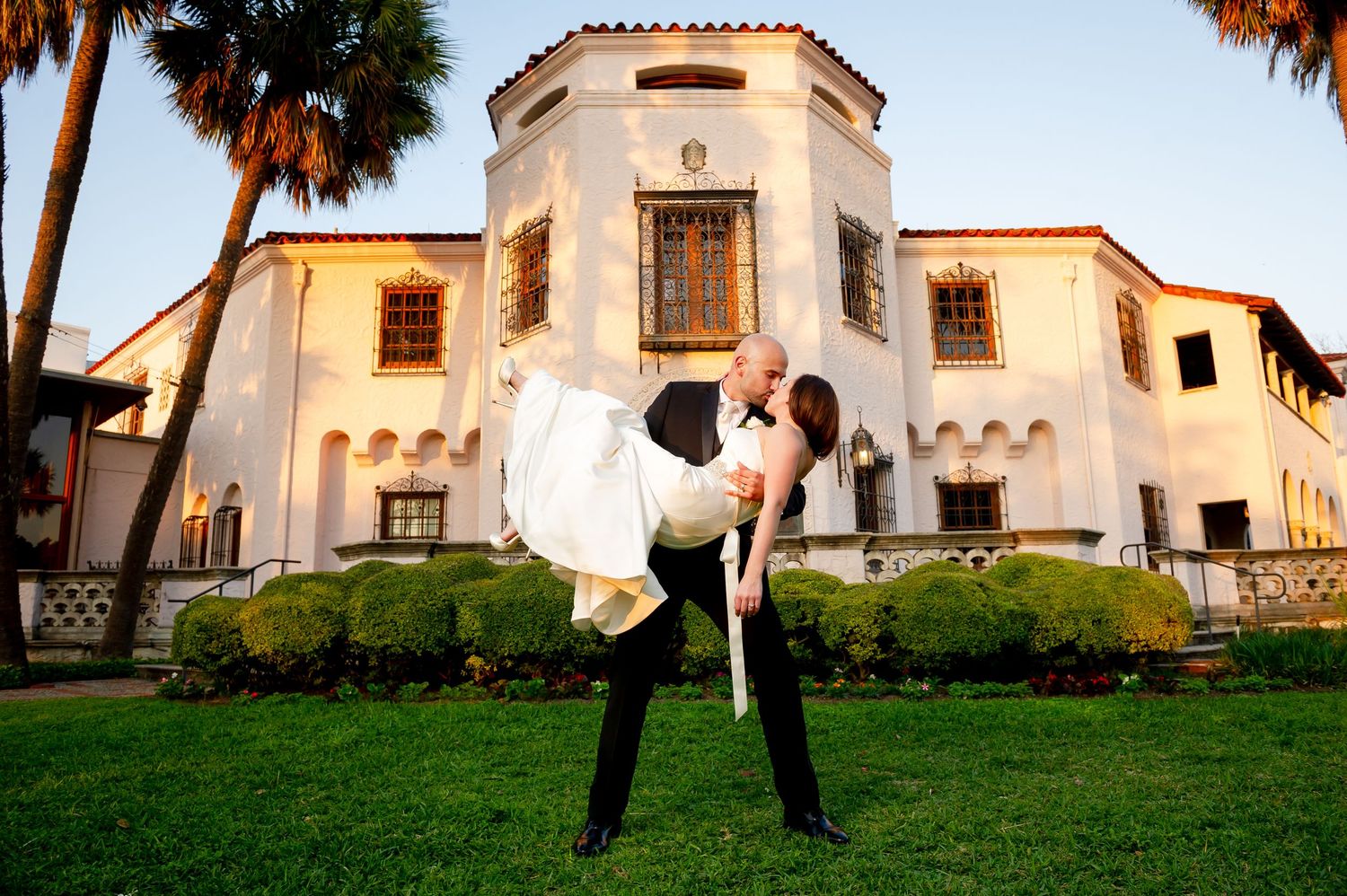 Romantic couple pose in front of a Spanish-style mansion with white walls and terracotta roof at dusk.