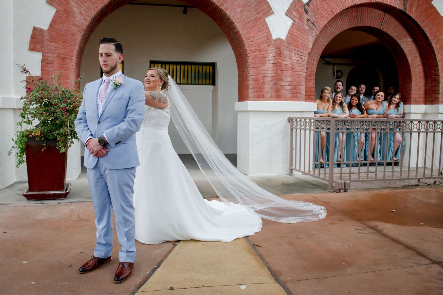 A couple in wedding attire stands under brick arches while bridesmaids watch from above.