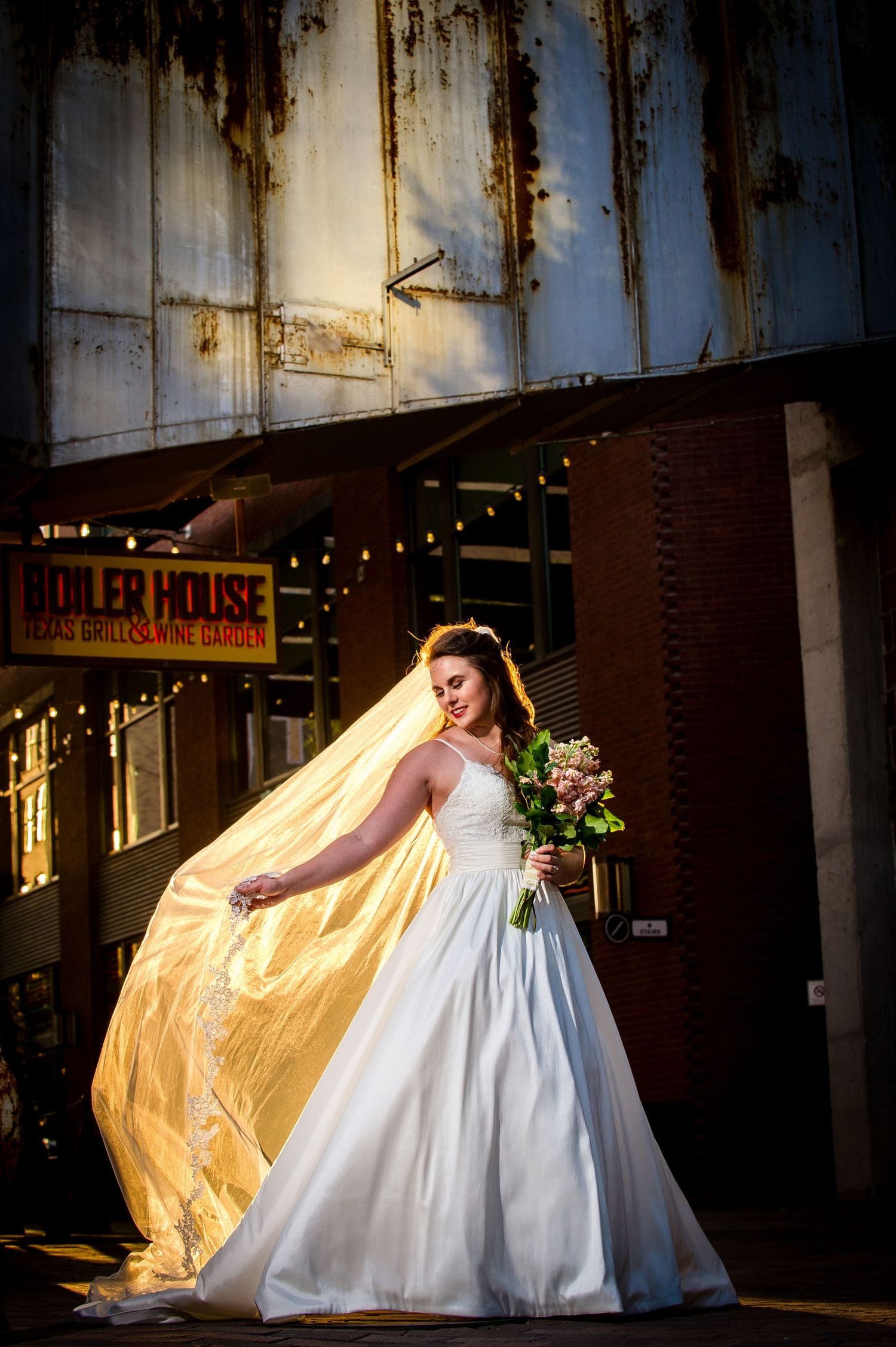 Dramatic bridal portrait with flowing veil against rustic brick building at sunset.