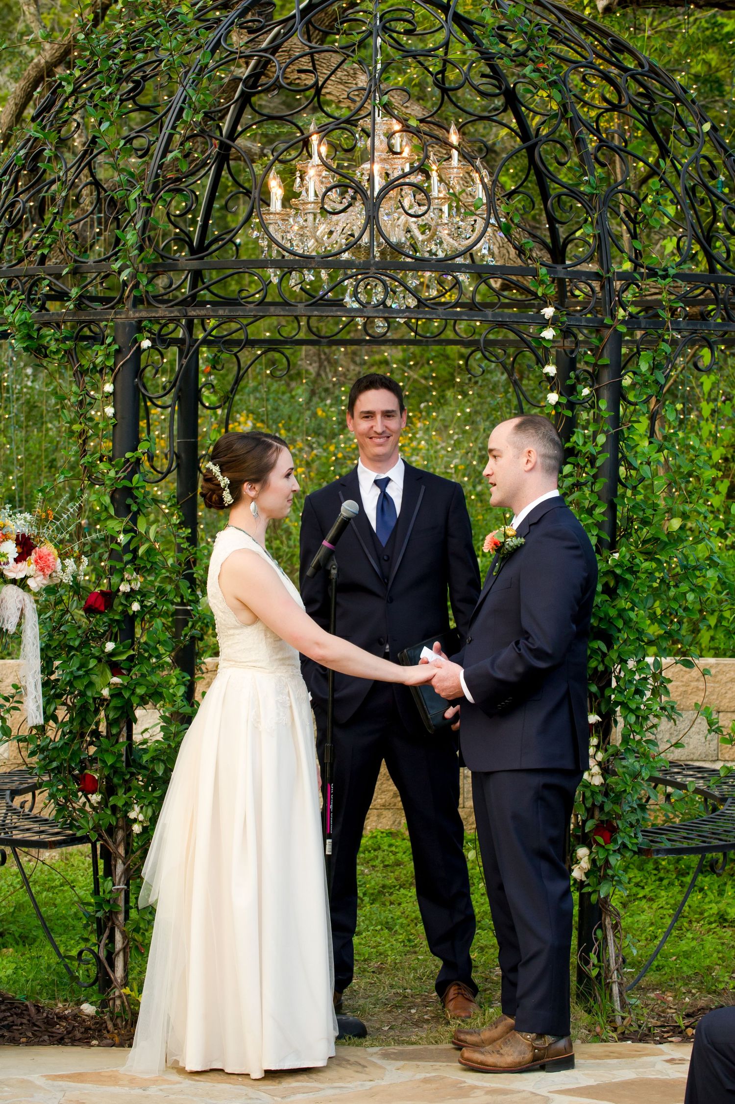 Outdoor wedding ceremony taking place under a lush green garden arbor with decorative ironwork.