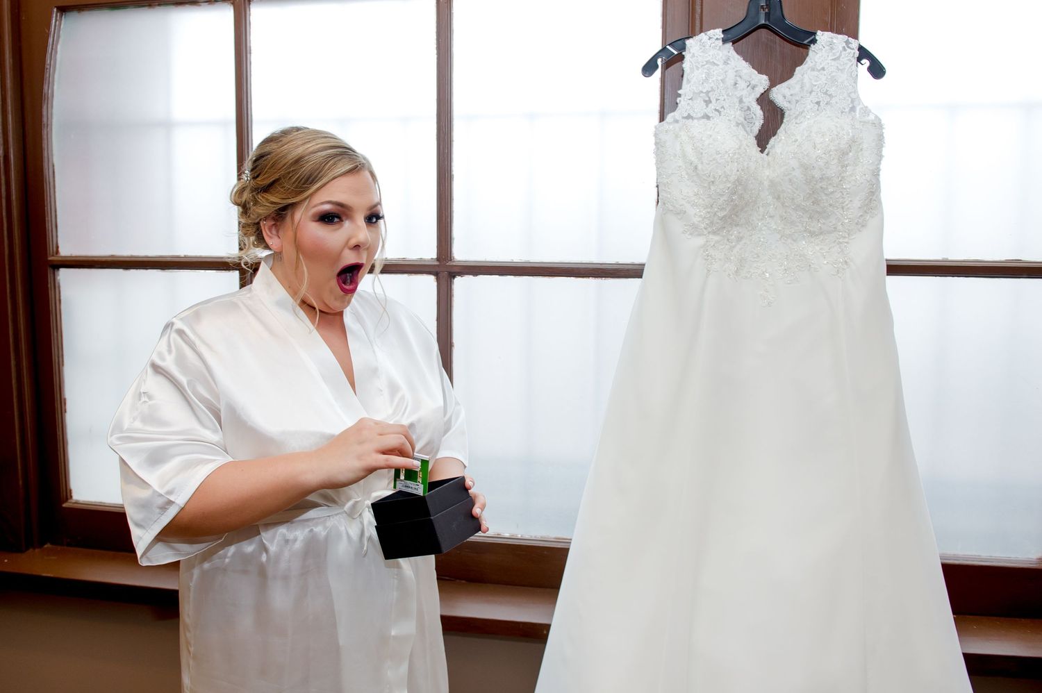 Wedding dress hanging in window while preparations take place in background.