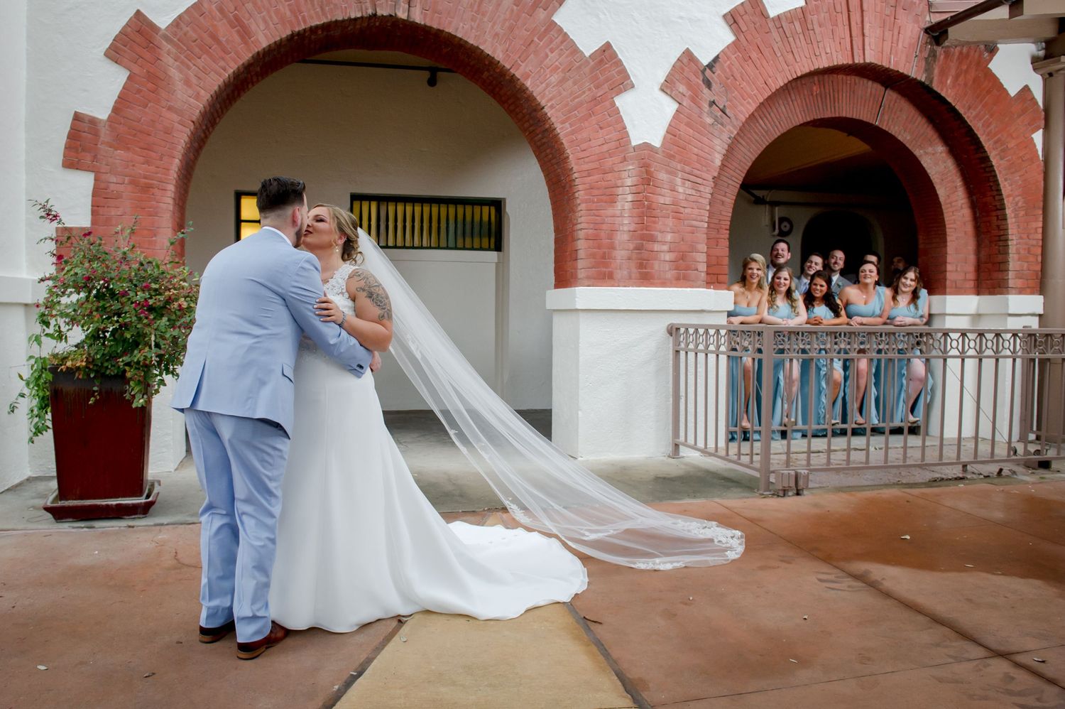 A couple shares their first look moment under brick arches while bridesmaids watch from above.
