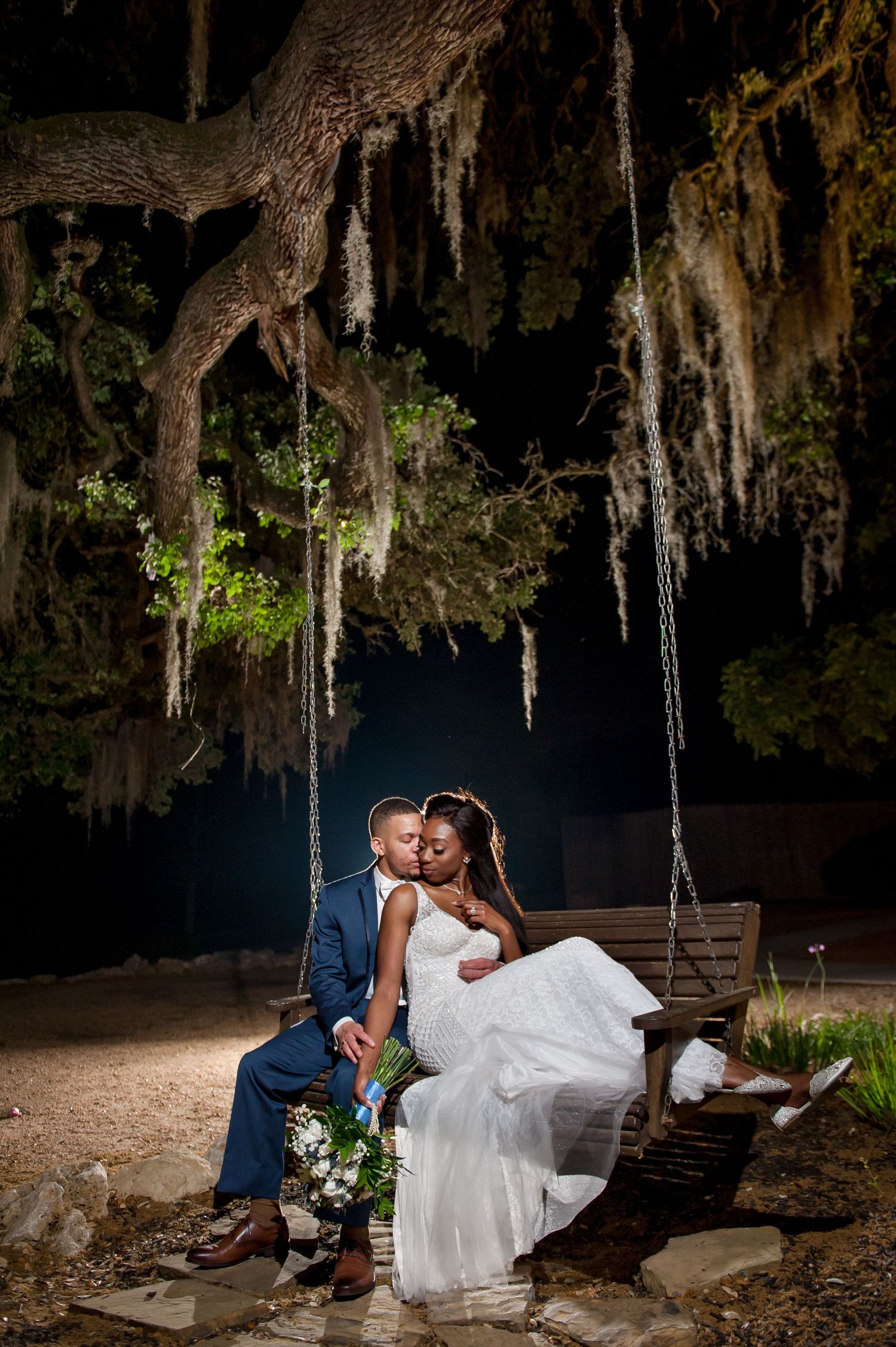 Couple shares an intimate moment on a swing under illuminated moss-covered trees at night.