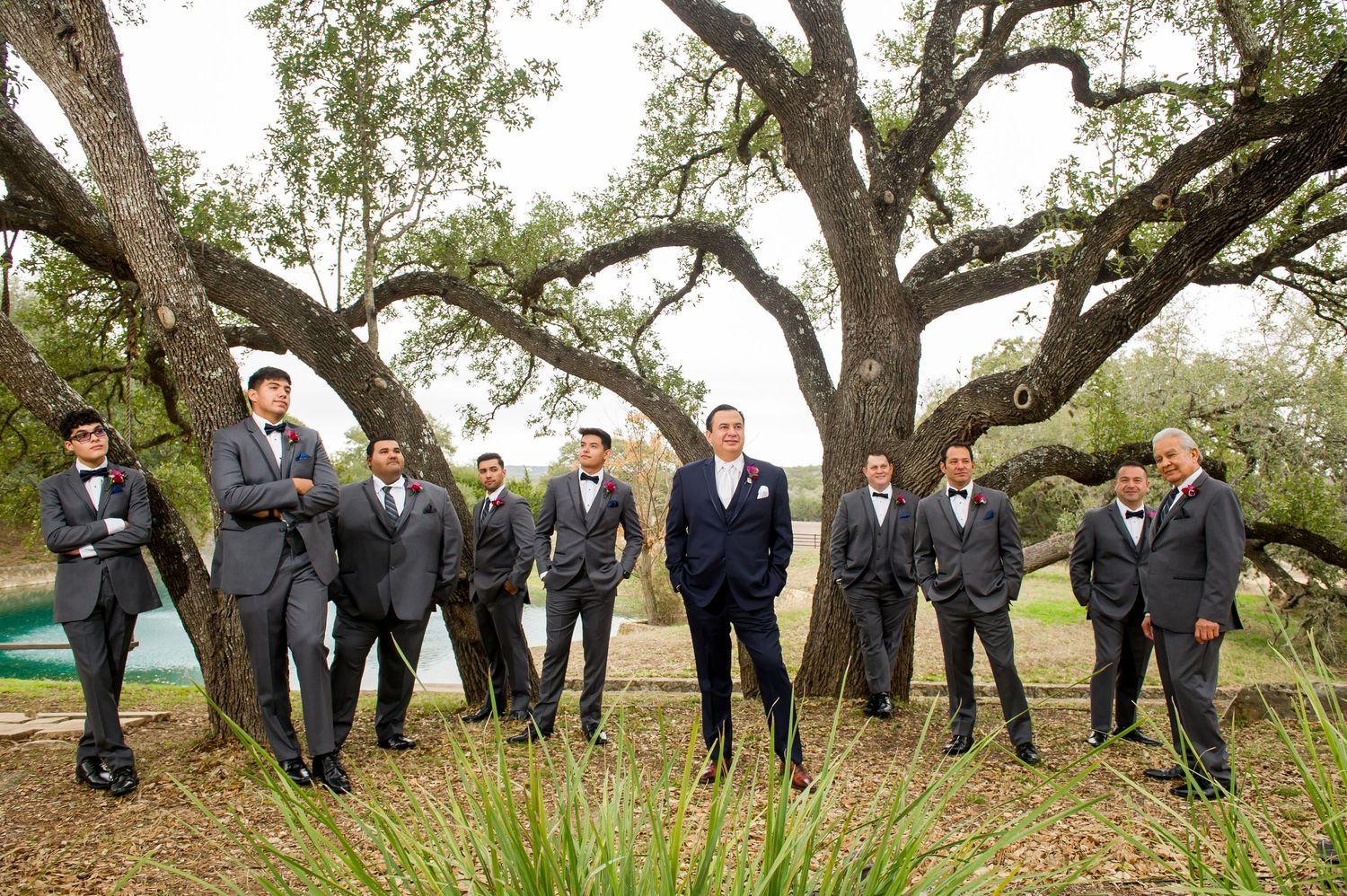 Groomsmen pose creatively under large oak trees for fun wedding party photo.