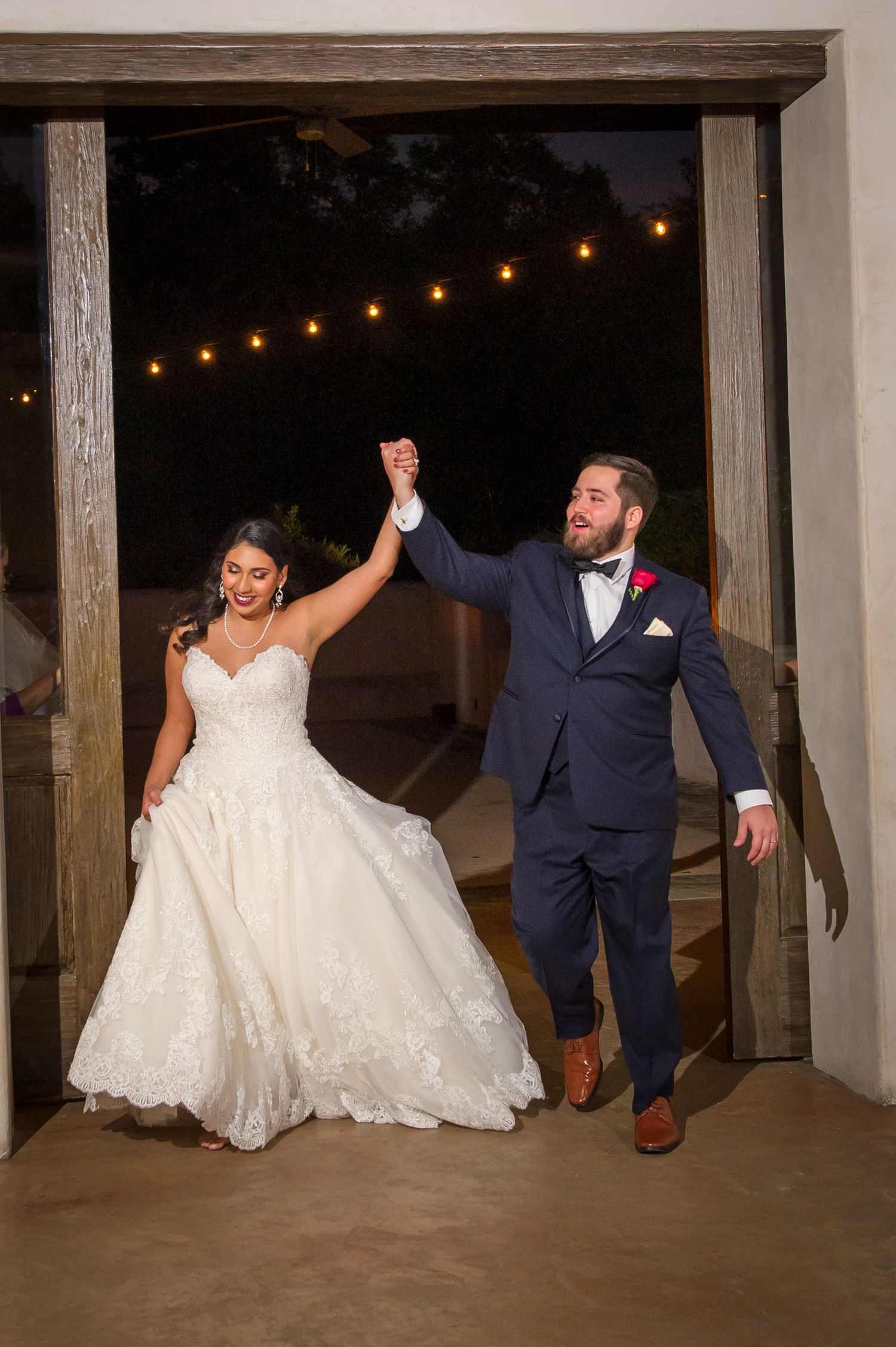 Wedding party celebrates with joyful dance moves in rustic venue with string lights.