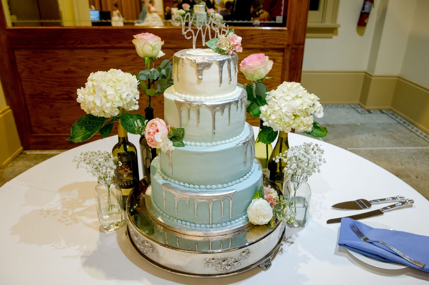 Elegant four-tier wedding cake decorated with silver drips and fresh flowers on white tablecloth.
