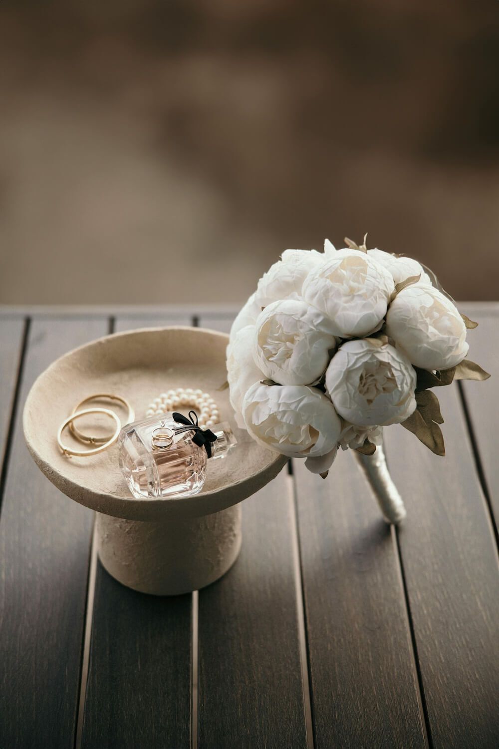 Wedding rings displayed on a cream-colored dish with delicate white peonies arranged on dark wooden planks.
