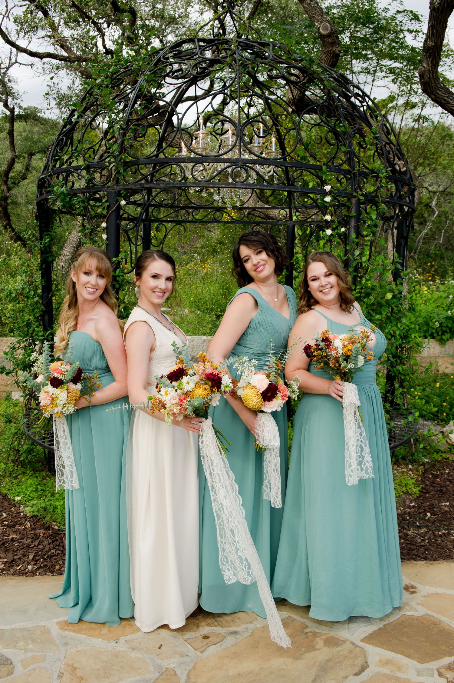 Wedding party wearing sage green dresses and white lace details pose with colorful bouquets in an outdoor garden setting.