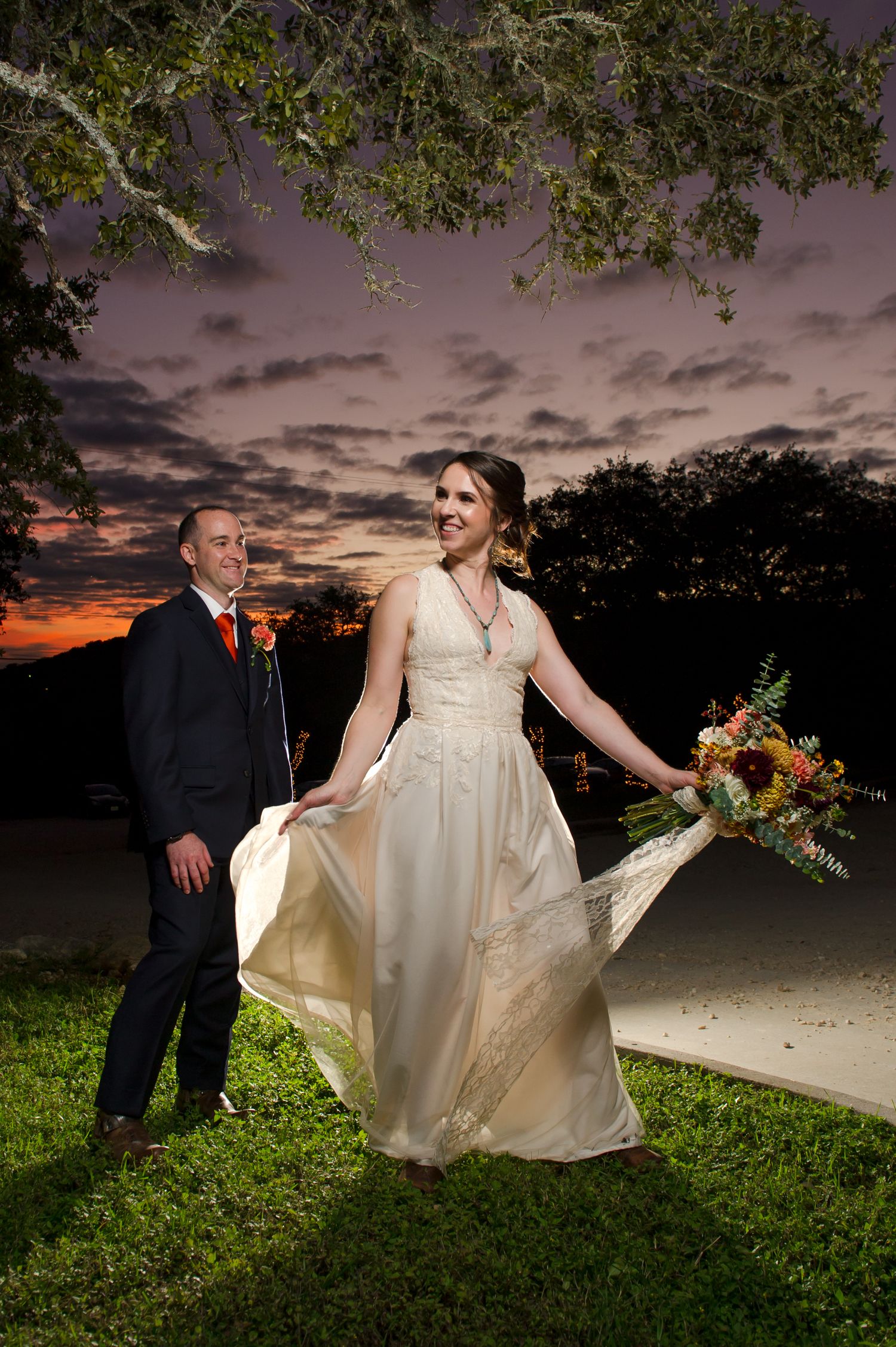 Wedding couple poses playfully on grass with dramatic evening sky backdrop.