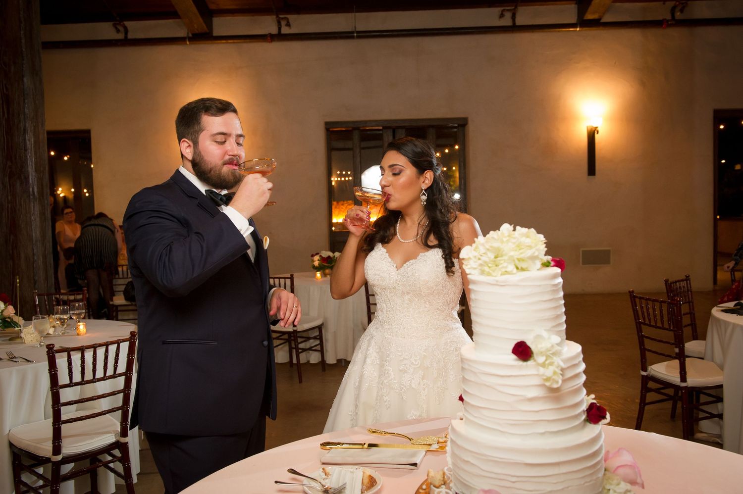 A couple shares an intimate moment while cutting their white tiered wedding cake at a reception.