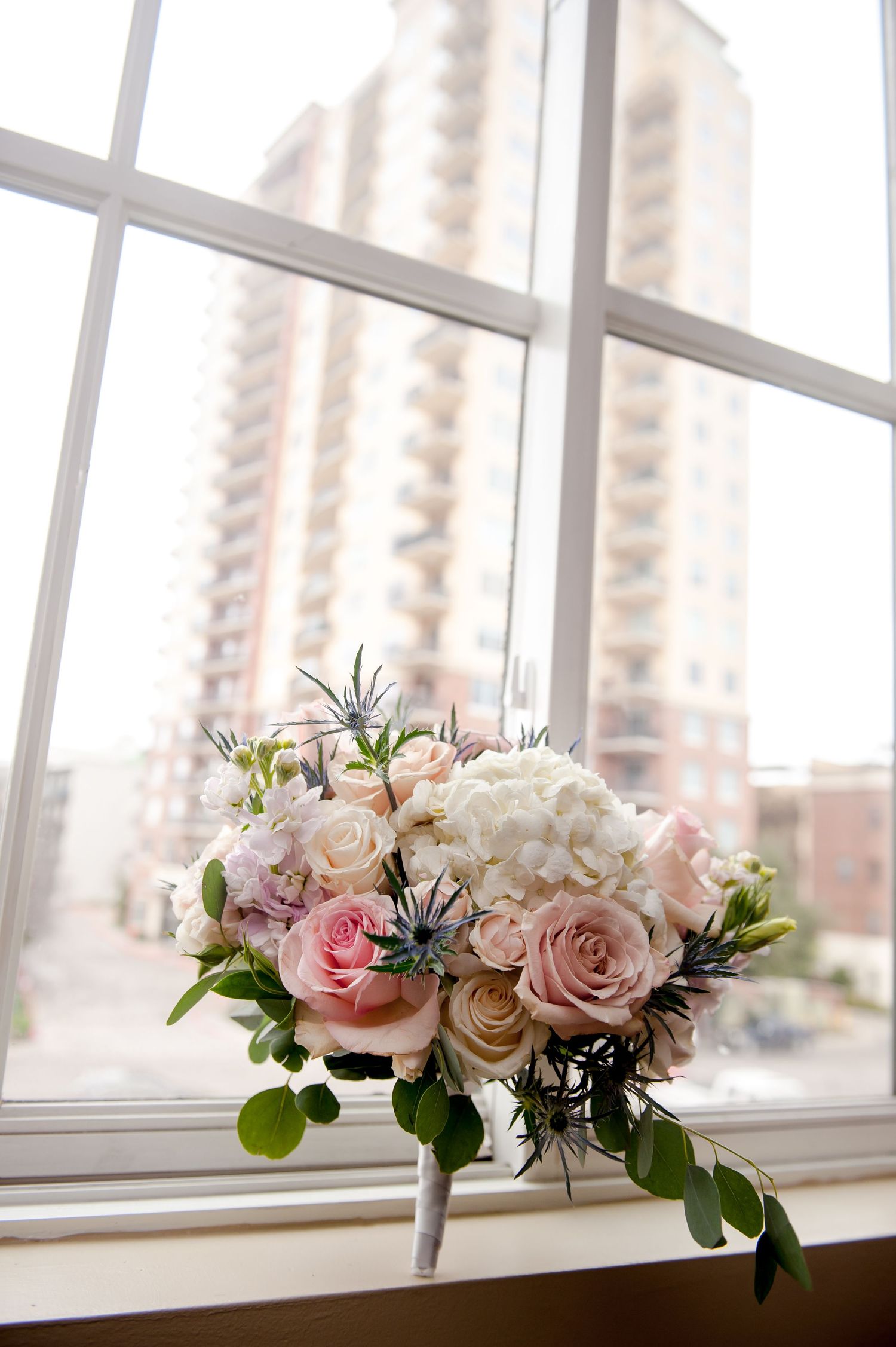 Delicate pink and white rose bouquet arrangement on windowsill with city view.