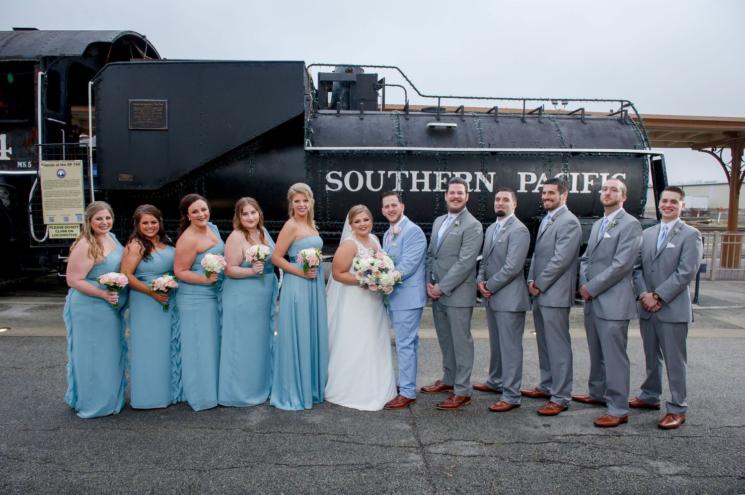 Wedding party lined up against vintage Southern Pacific train in blue dresses and gray suits.
