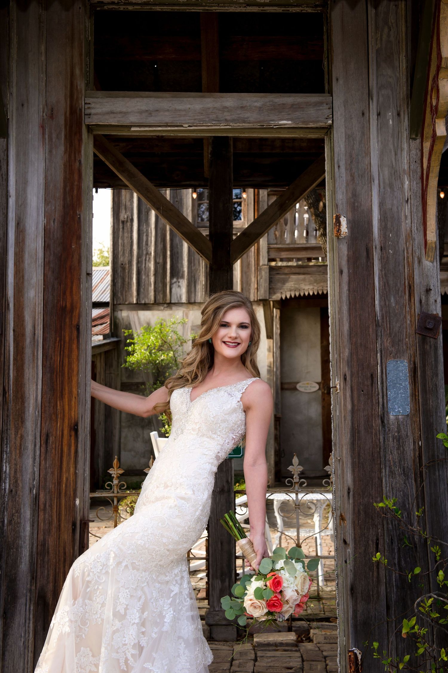 A white lace wedding dress photographed in a rustic wooden doorway with a bouquet of flowers.