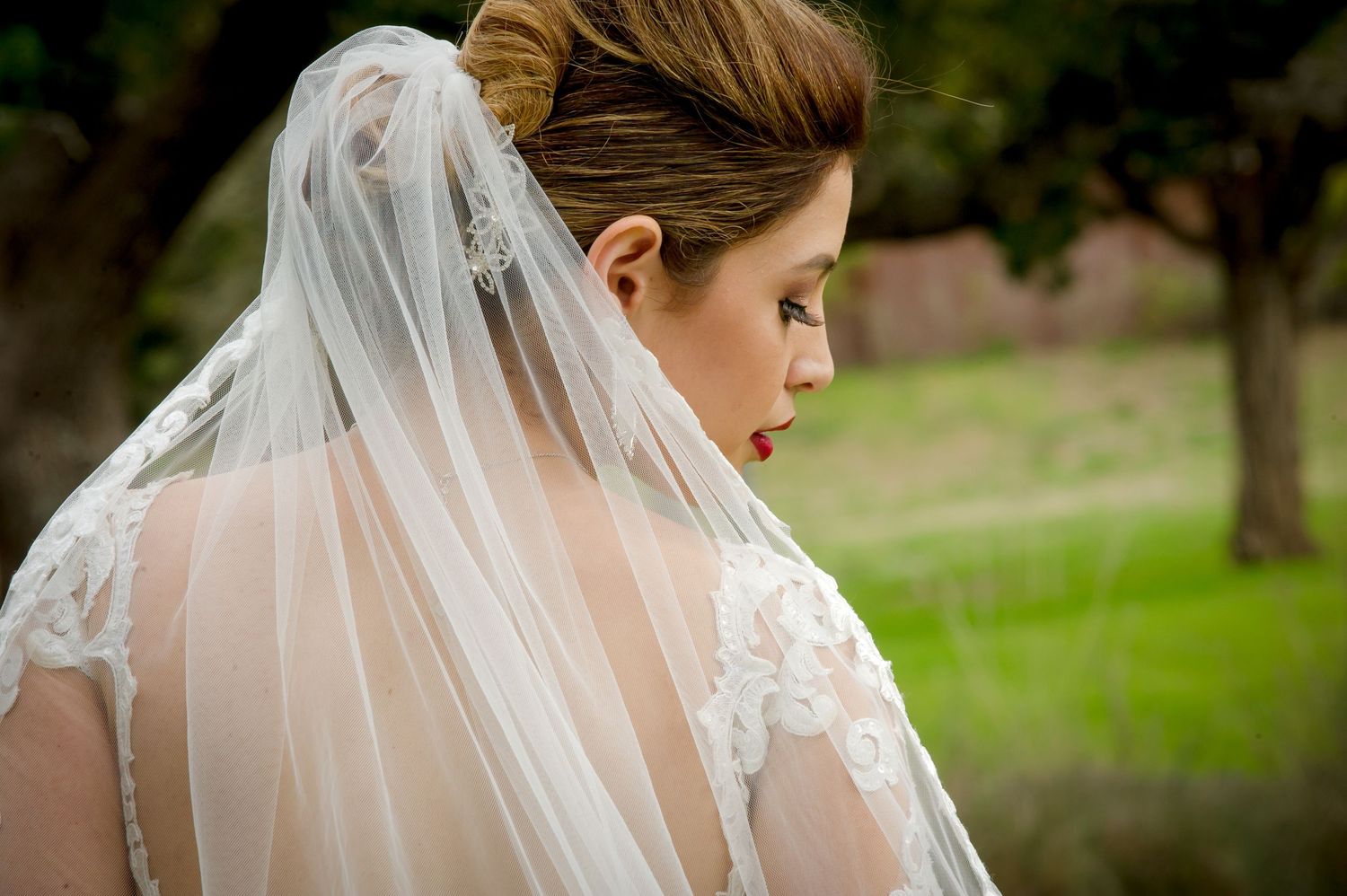 A bride in a delicate lace veil poses outdoors in a natural garden setting.