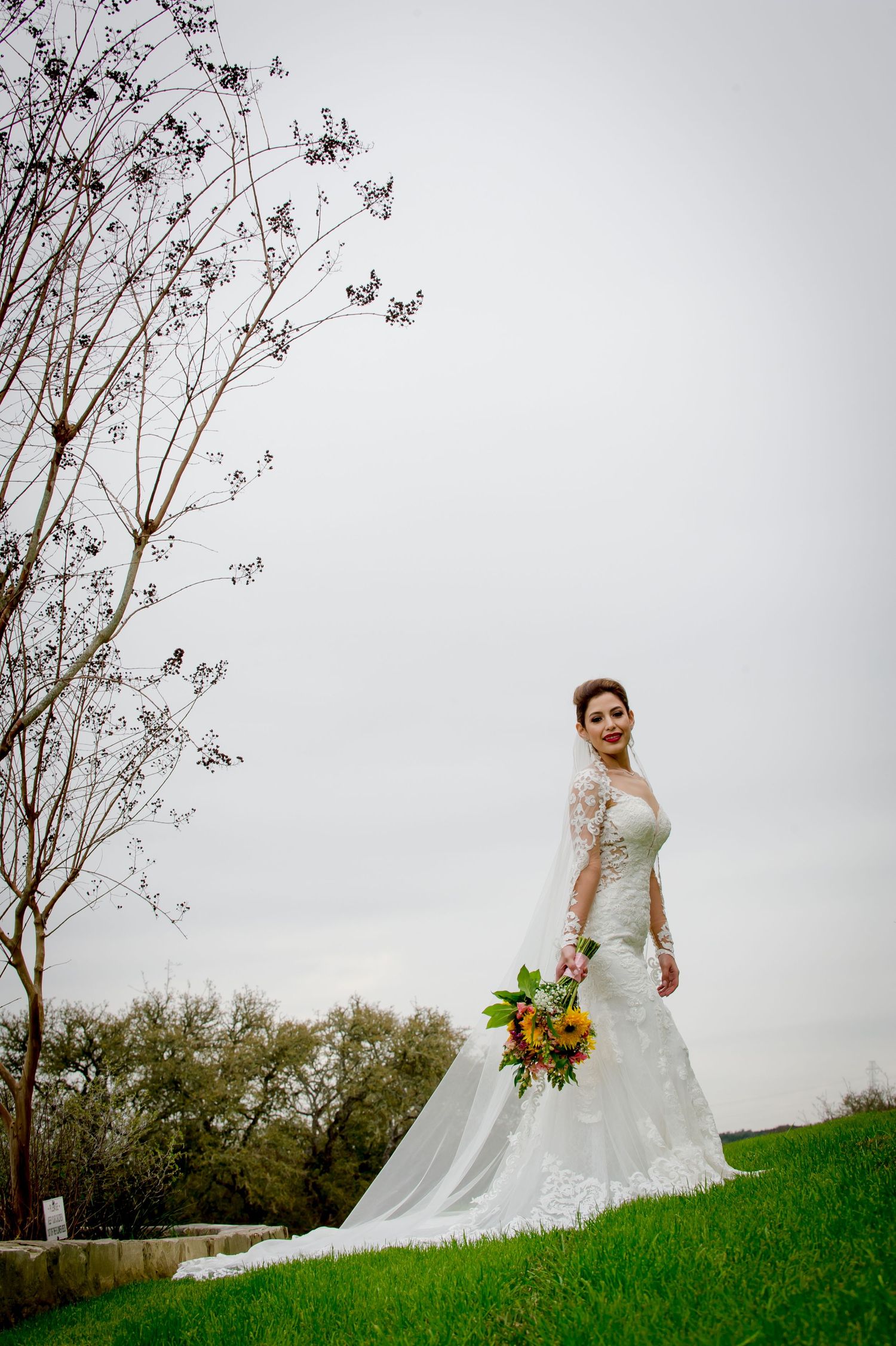 A bride in a lace white wedding dress stands on a grassy hill holding a vibrant sunflower bouquet against an overcast sky.