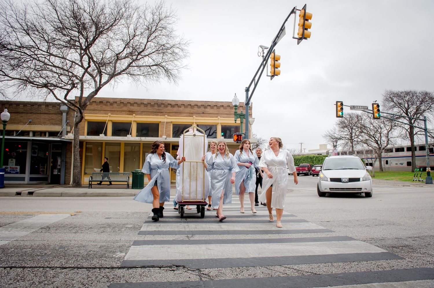 Wedding party crosses city street with traffic signals and bare trees in background.