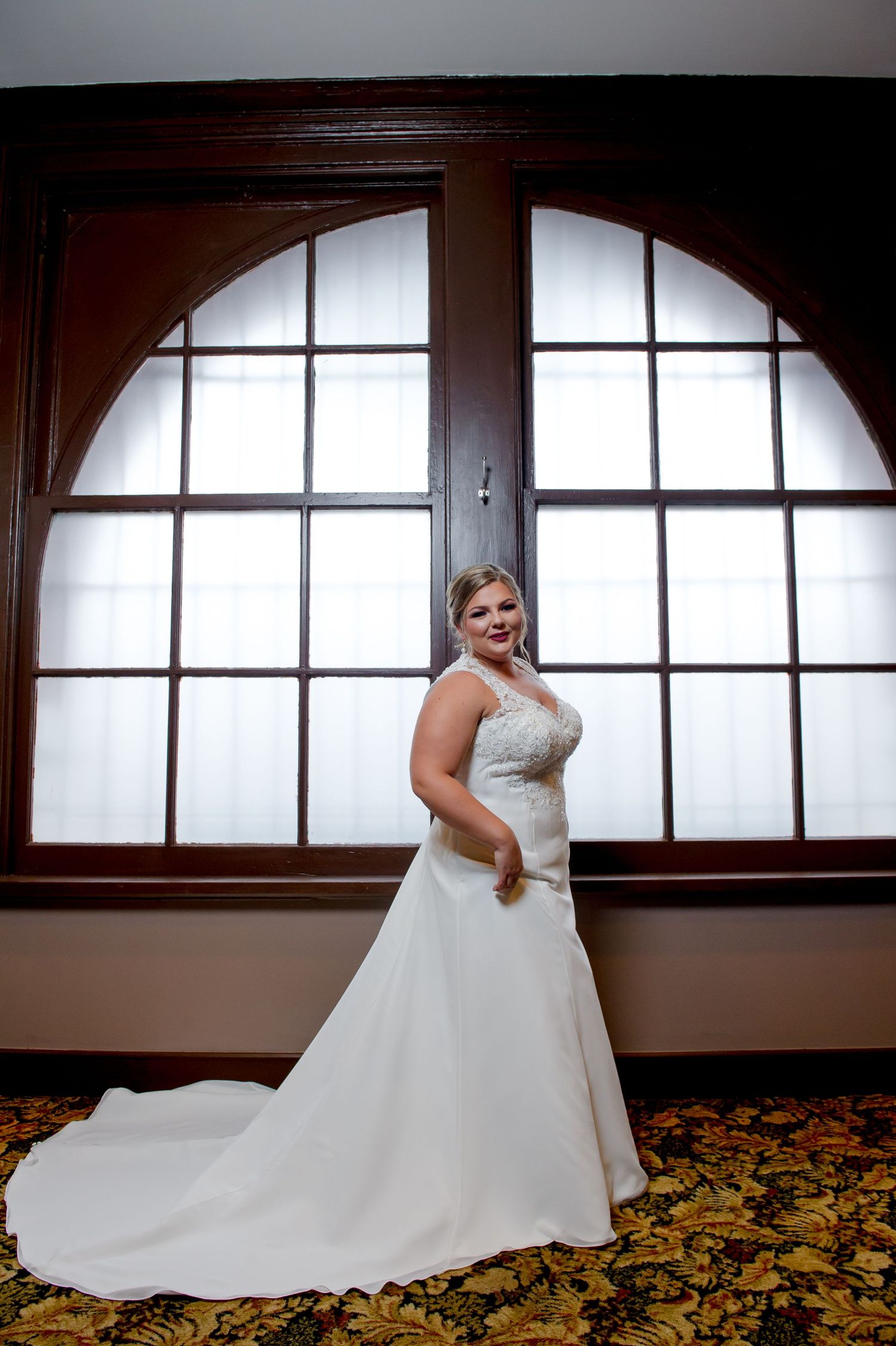 A bride in a flowing white gown poses in front of a large arched window.