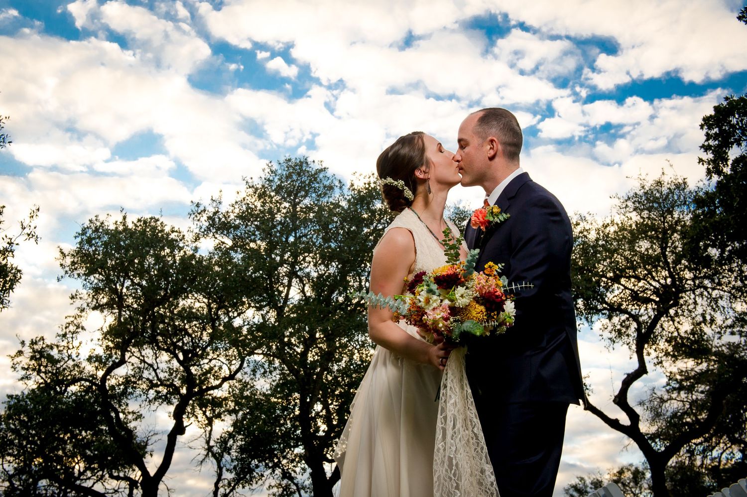 A couple shares a romantic kiss at sunset with silhouetted trees and dramatic clouds in the background.