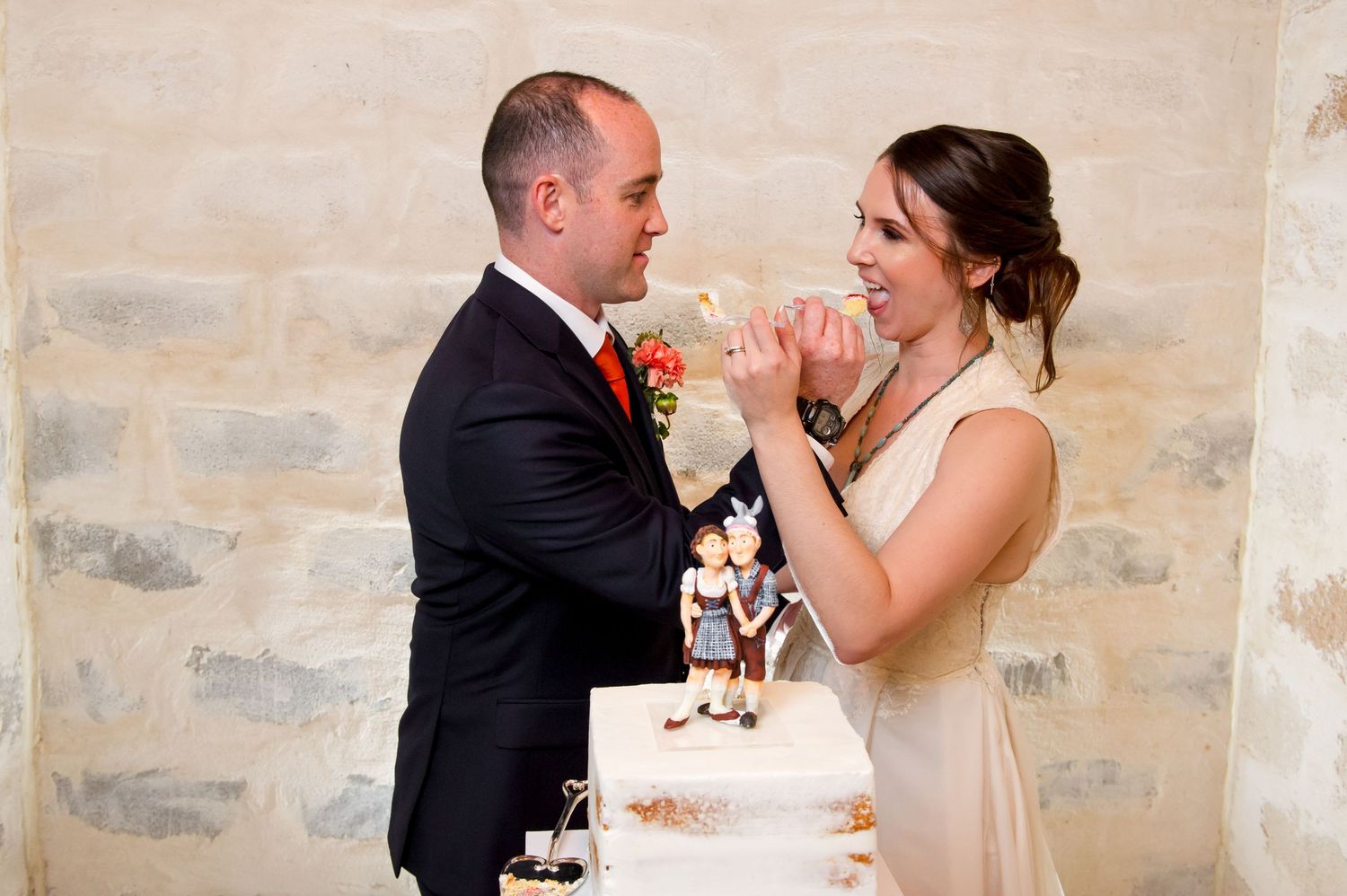 A couple shares a bite of wedding cake during their reception celebration against a stone wall backdrop.