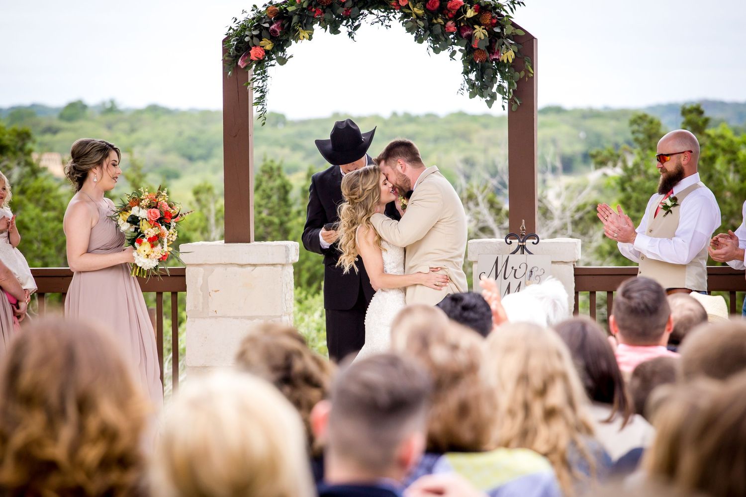 Outdoor wedding ceremony under floral arch with guests seated in rustic venue.