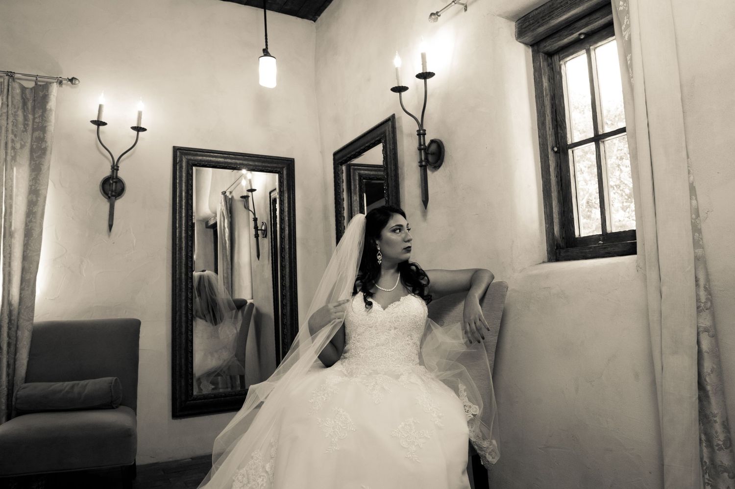 Black and white image of bride in full ballgown seated in vintage room with wall sconces.