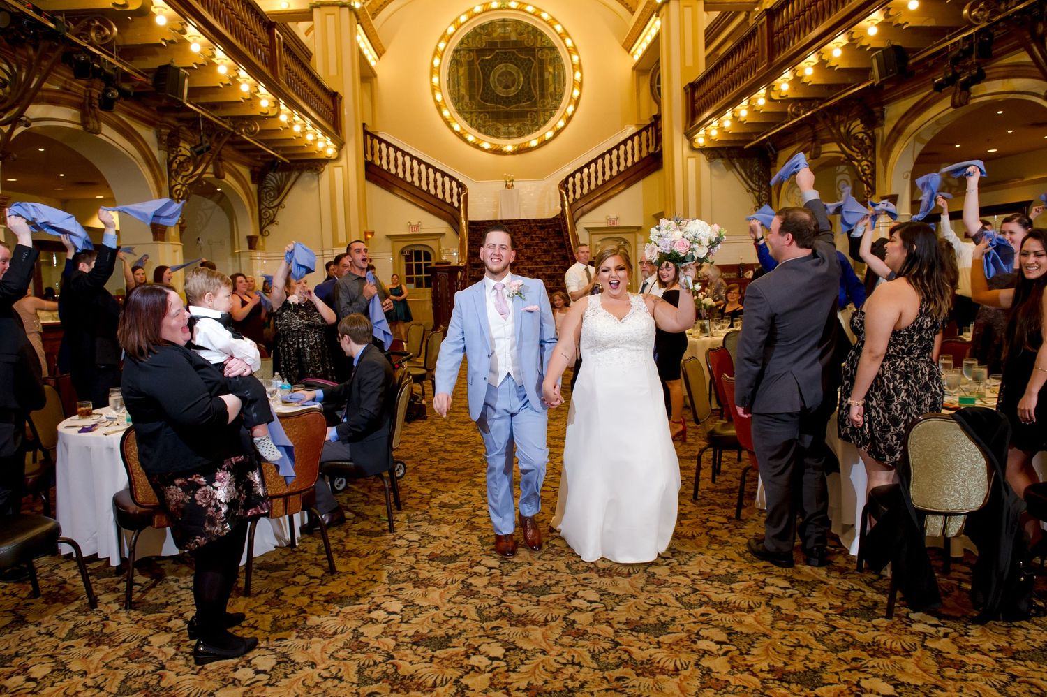 Wedding couple walks down grand ballroom aisle while guests cheer.