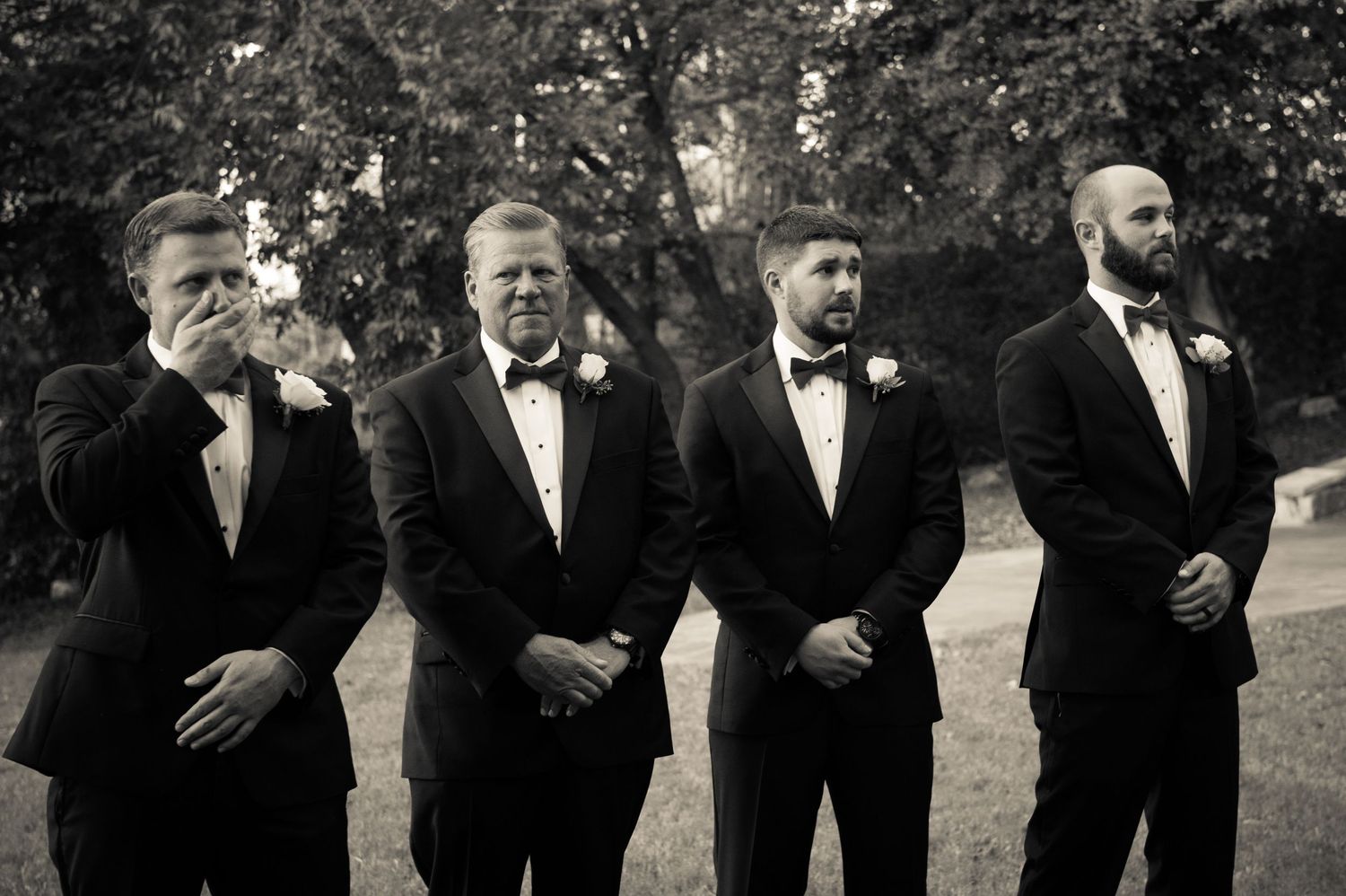 Groomsmen in black tuxedos line up for formal wedding photos outdoors.