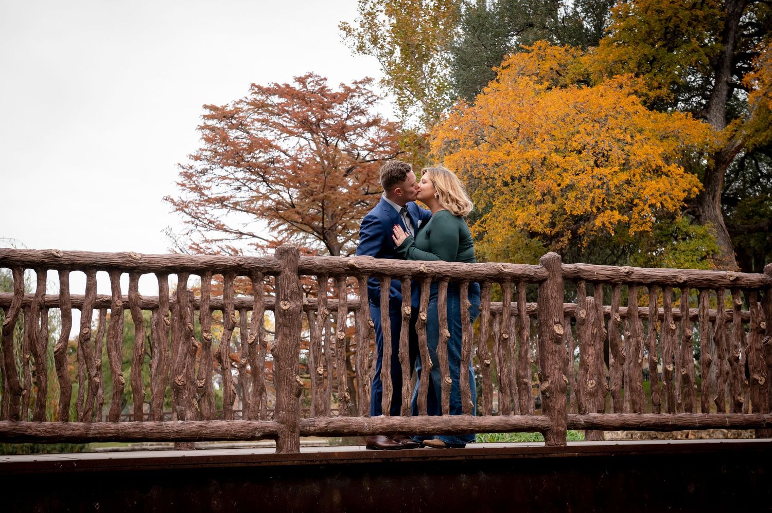 Couple embraces on wooden bridge surrounded by vibrant autumn foliage in romantic outdoor setting.