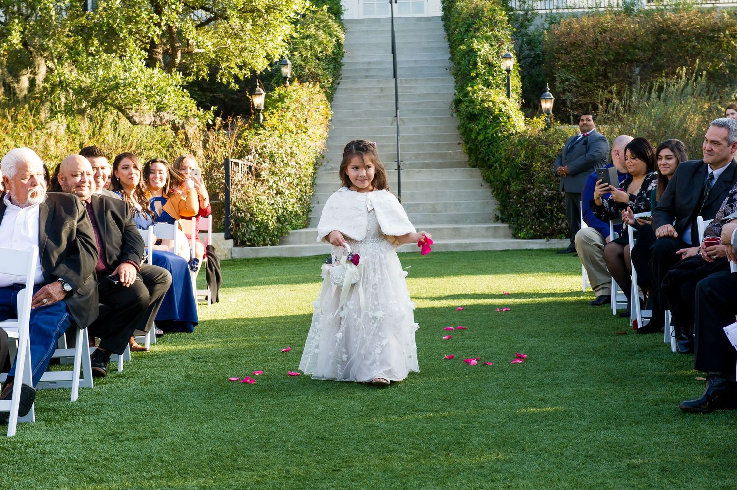 Outdoor garden wedding ceremony with guests seated on white chairs on manicured lawn.