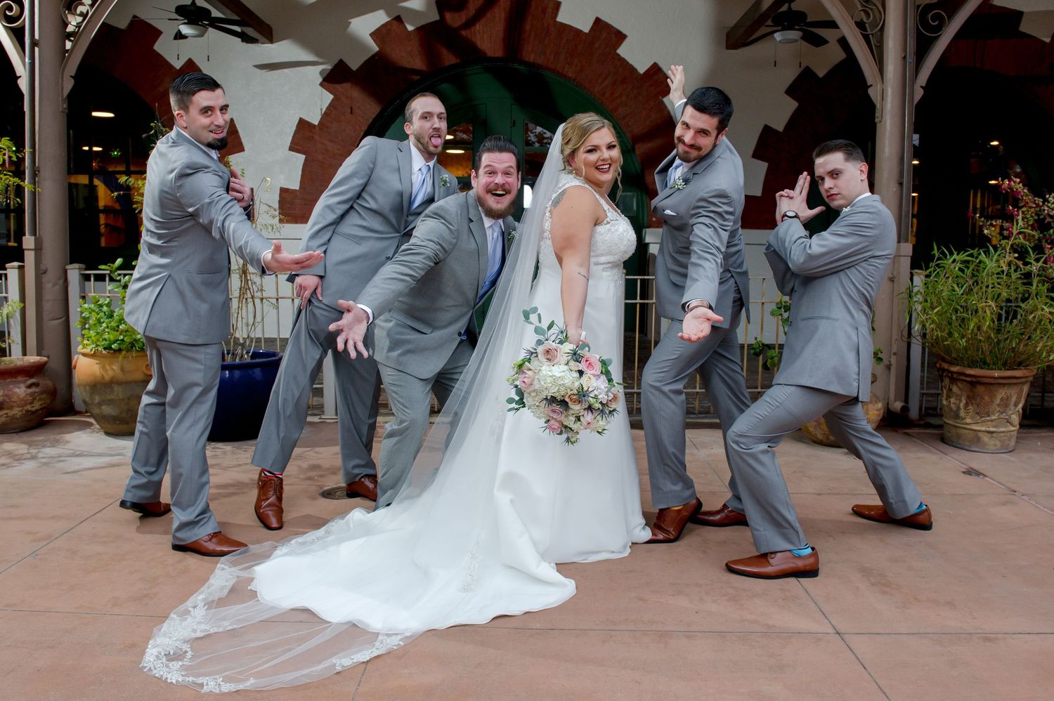 Wedding party in gray suits and white dress dance and pose playfully on outdoor patio.