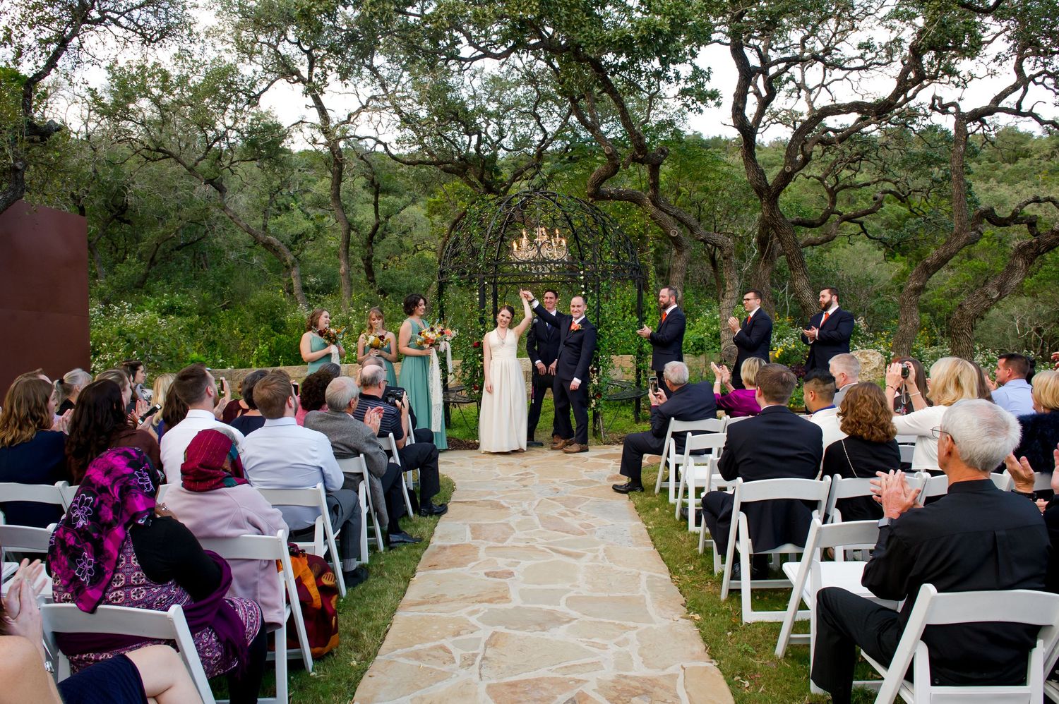Stone patio outdoor wedding venue with white chairs arranged in rows under oak trees.