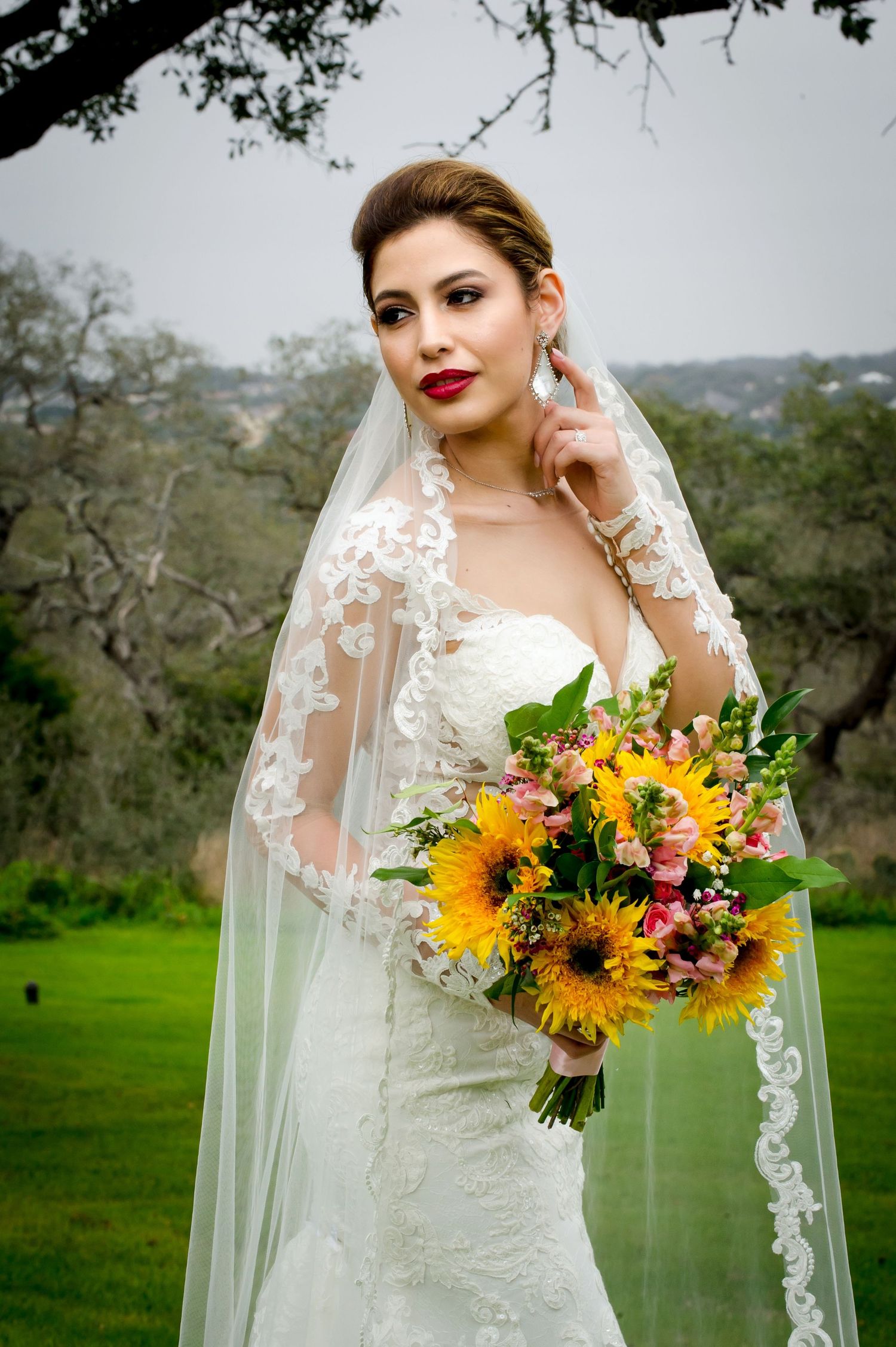 A bride in a white lace gown holds a vibrant sunflower bouquet while posing outdoors in a natural setting.