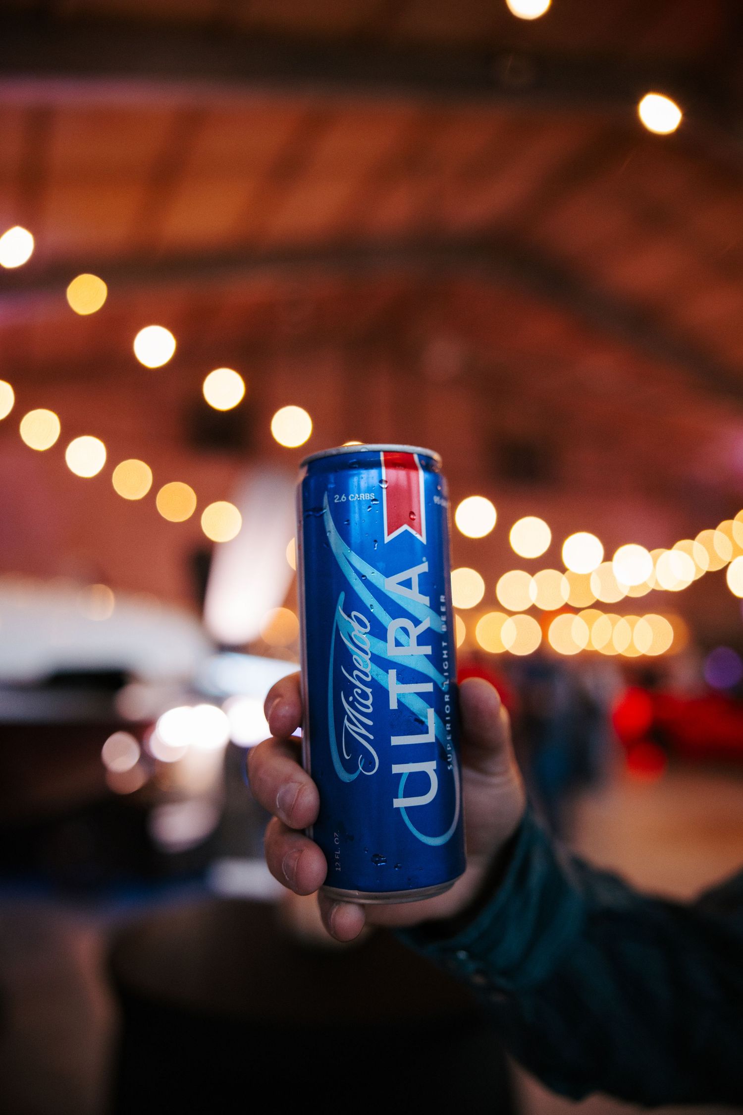 Hand holding a blue beer can in front of bokeh lights at an event