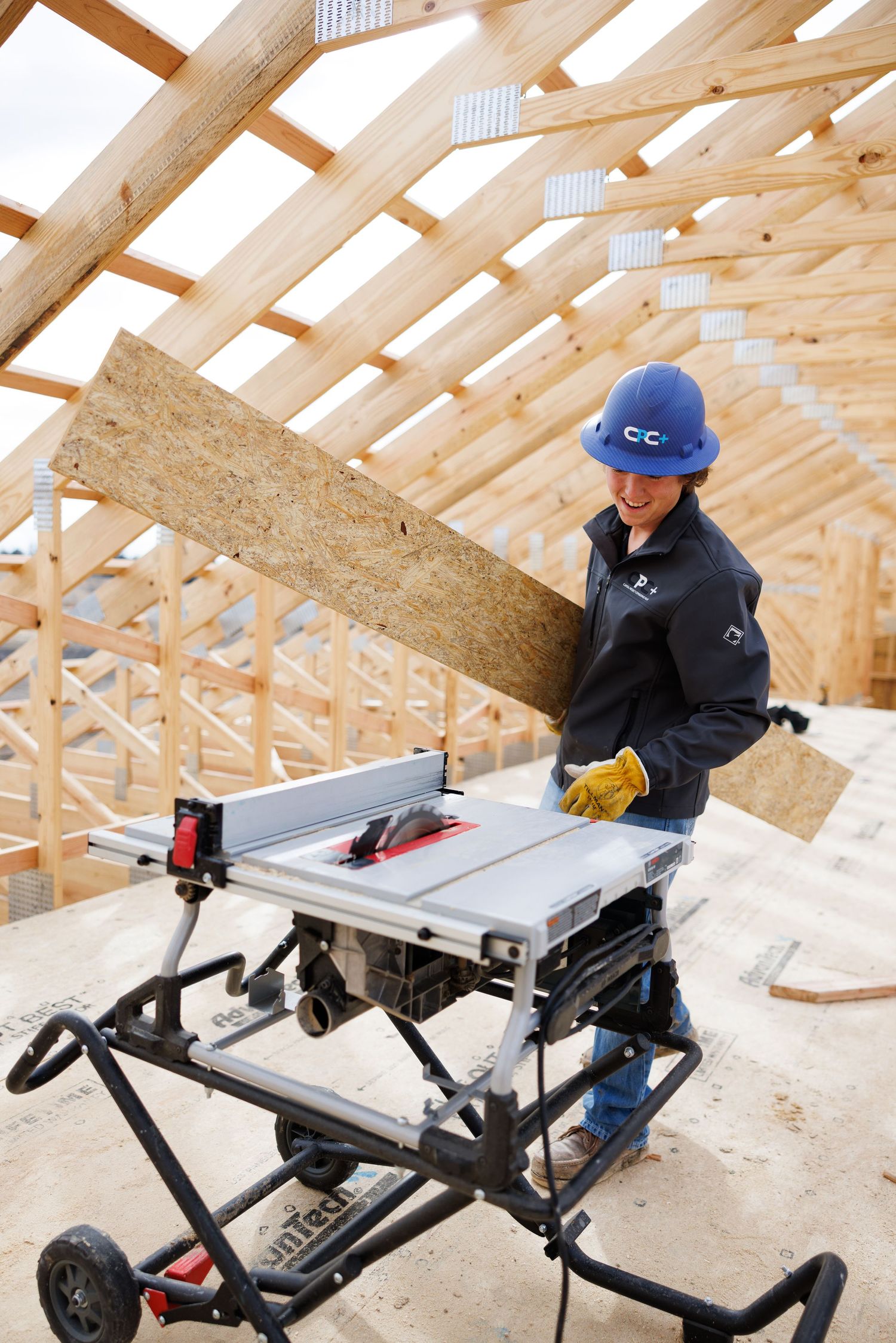 Image of home builder wearing a blue hardhat and gloves holding a piece of plywood next to a saw.