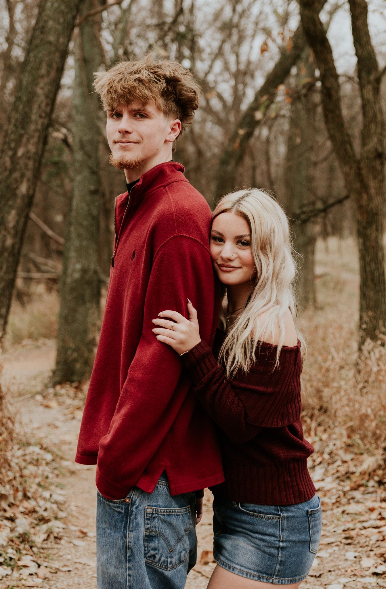 Young couple in matching burgundy outfits posing back-to-back on an autumn woodland path.
