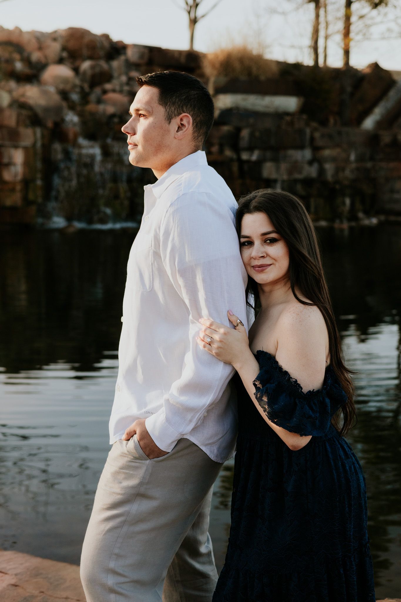 Woman in navy dress hugging man in white shirt from behind near scenic waterfall at golden hour.