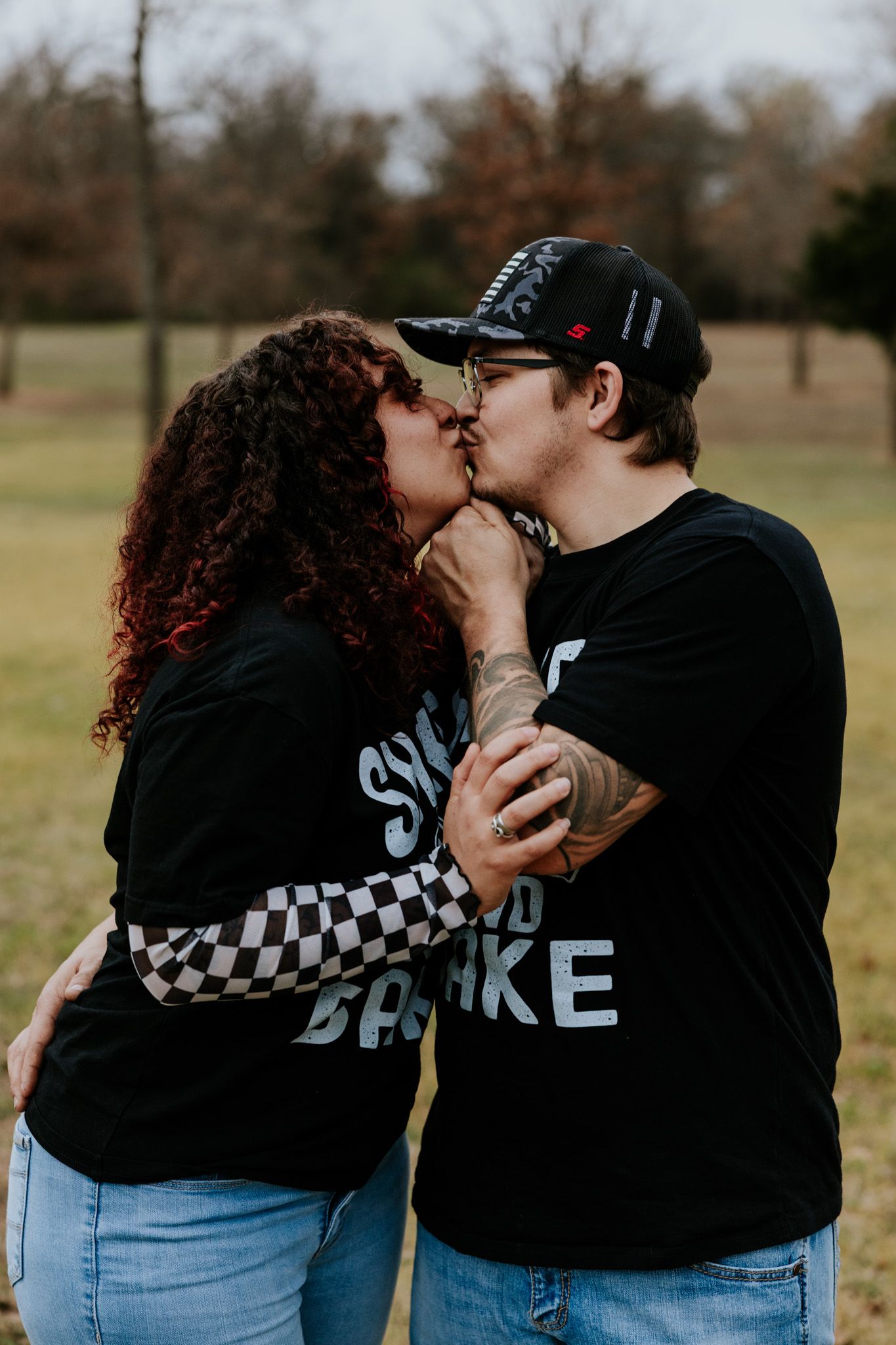 Couple sharing a kiss outdoors in a park, wearing matching Shake and Bake t-shirts and jeans.