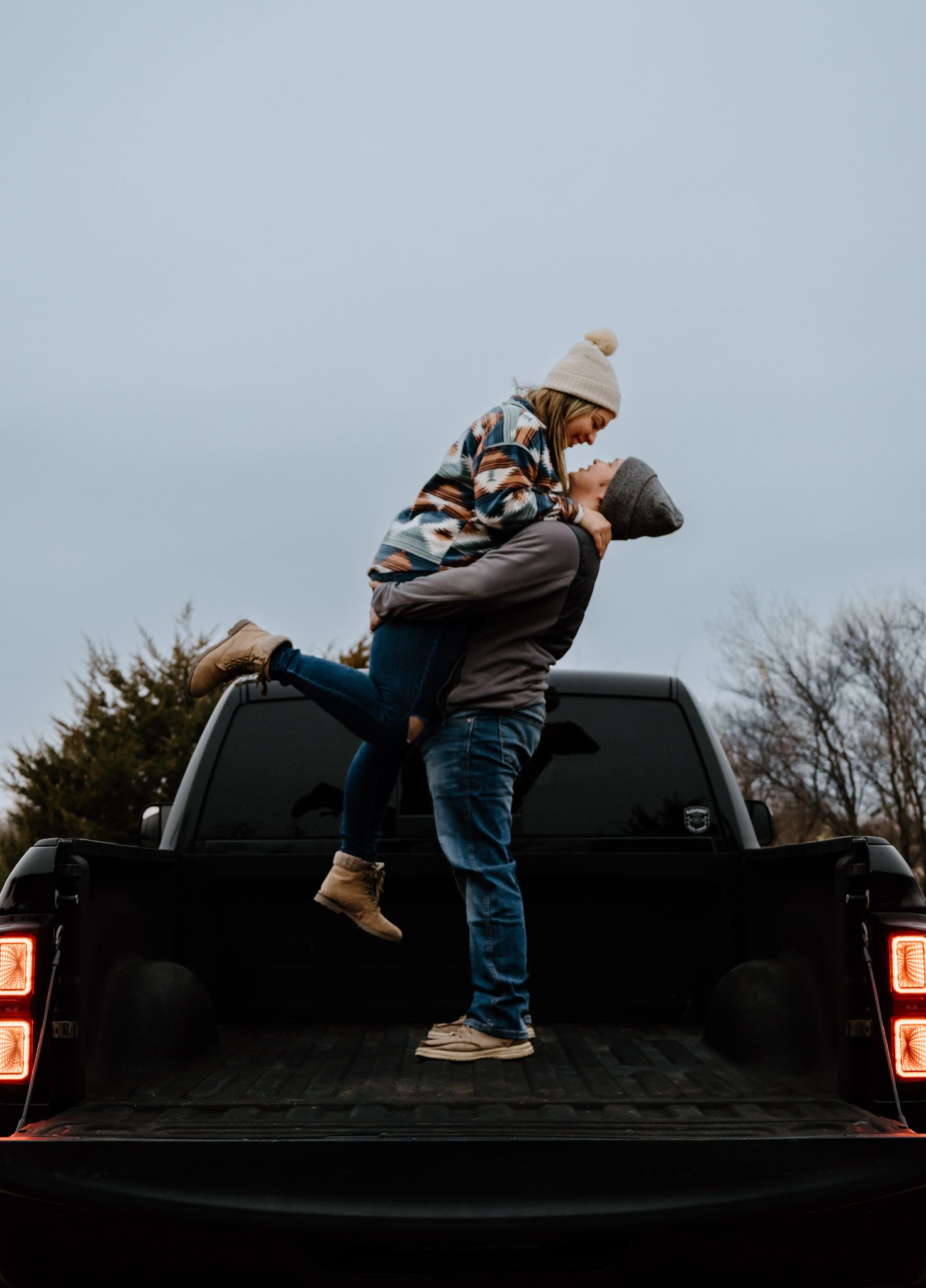 Romantic couple kissing as man lifts woman in truck bed against a moody winter sky.