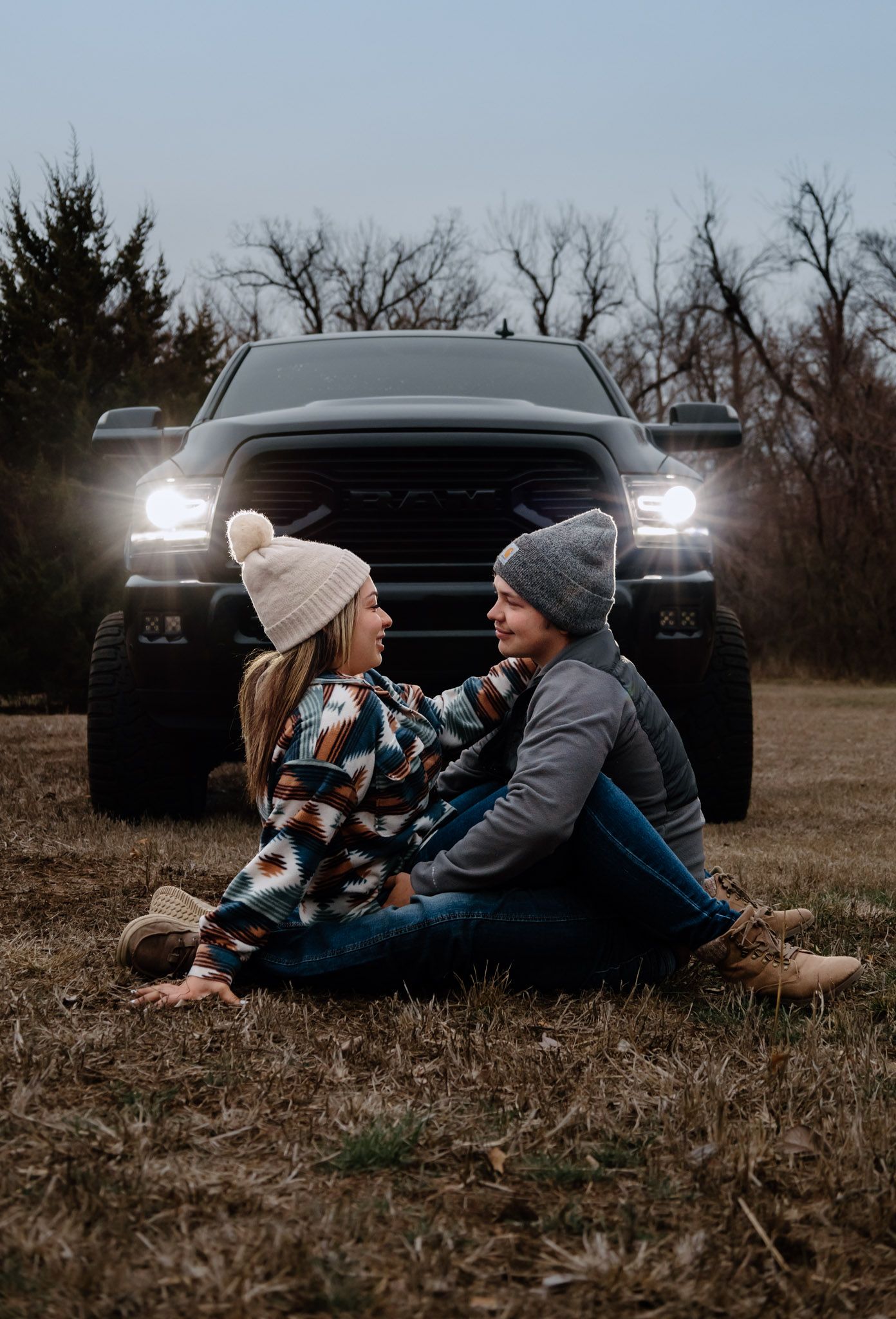 Couple sitting together on winter ground in front of a black RAM truck with headlights on.