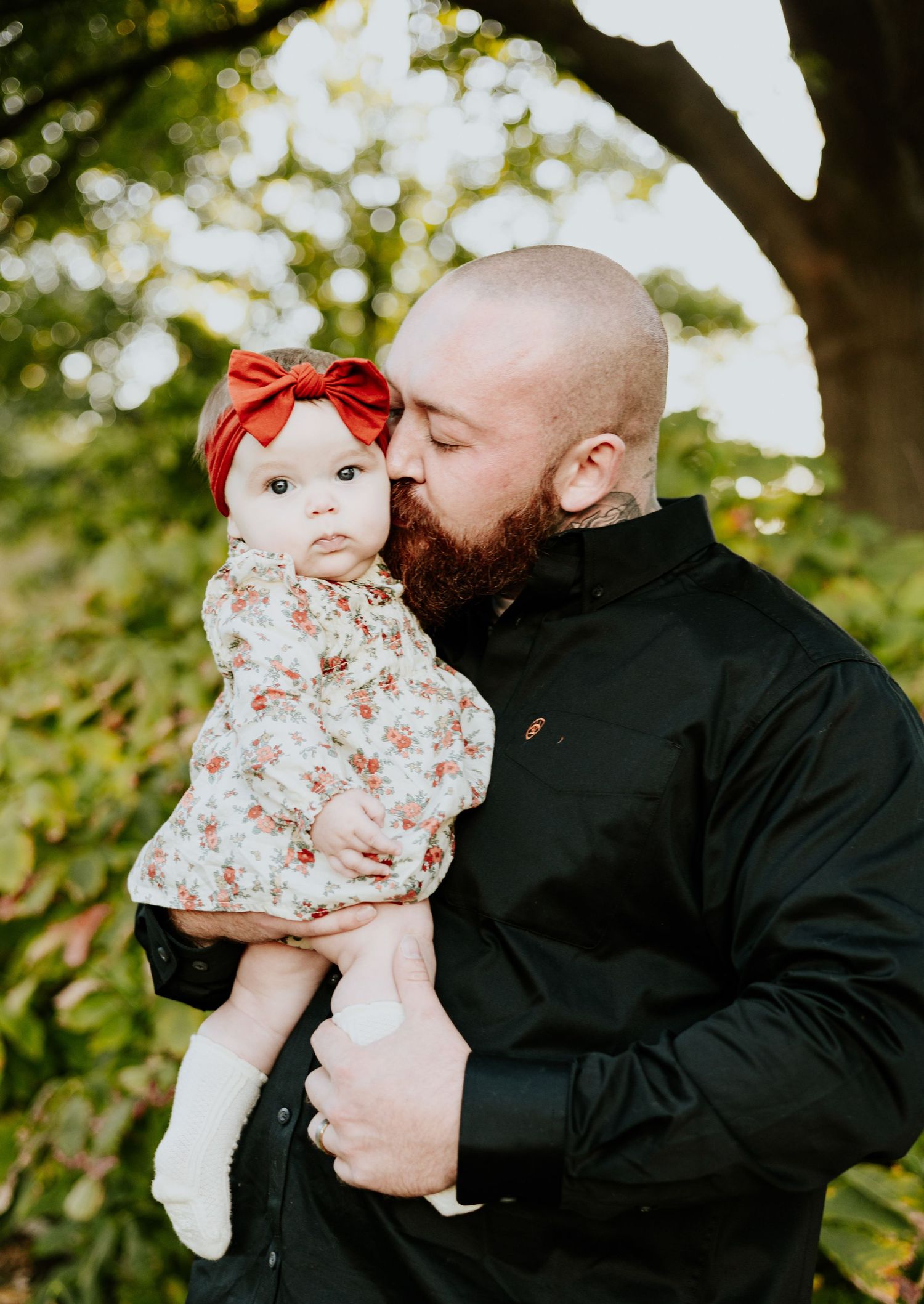 A bald bearded father in a black shirt tenderly kisses his infant daughter in a floral outfit with a red bow.