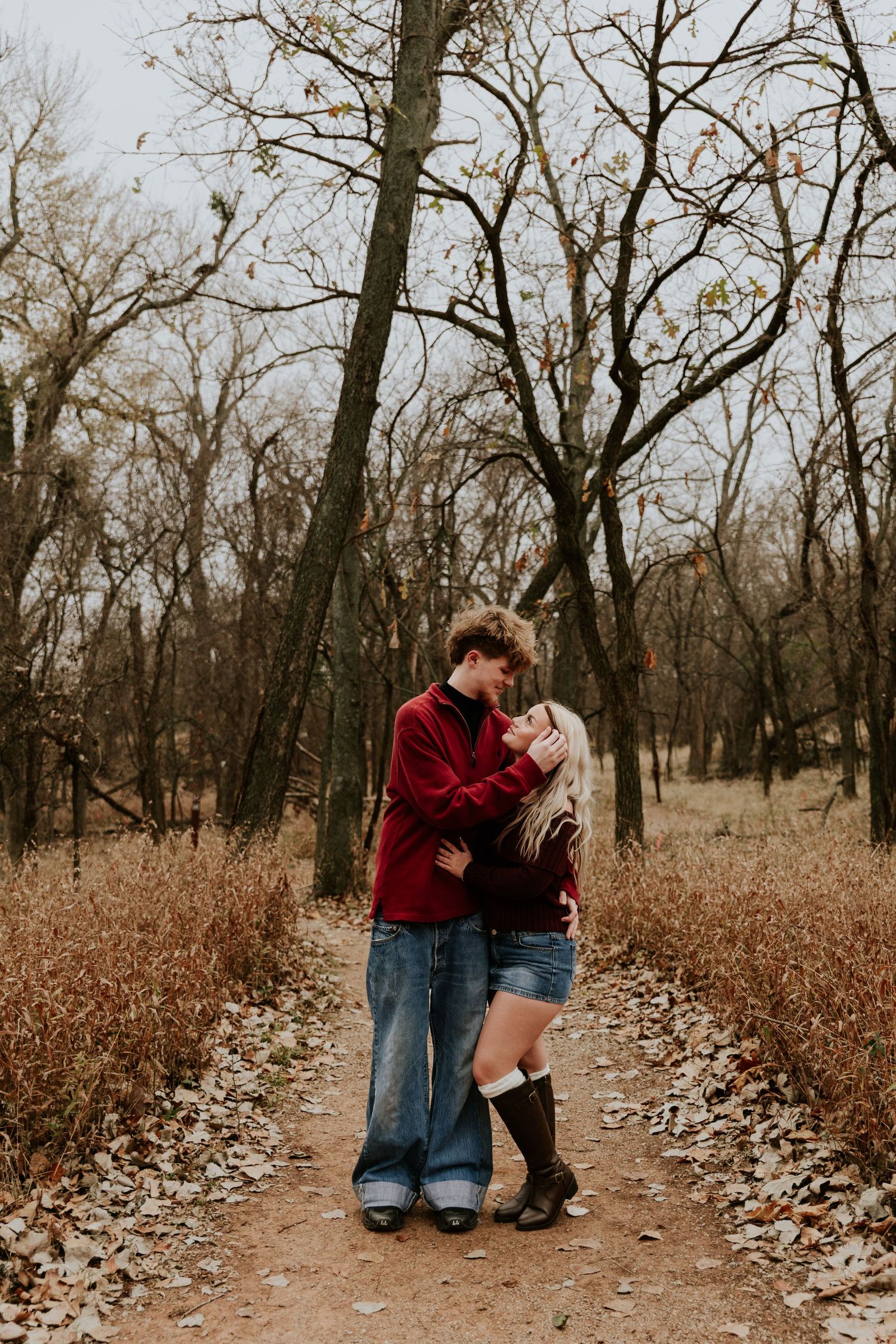 Young couple embracing and kissing on a leaf-covered forest path surrounded by bare autumn trees.