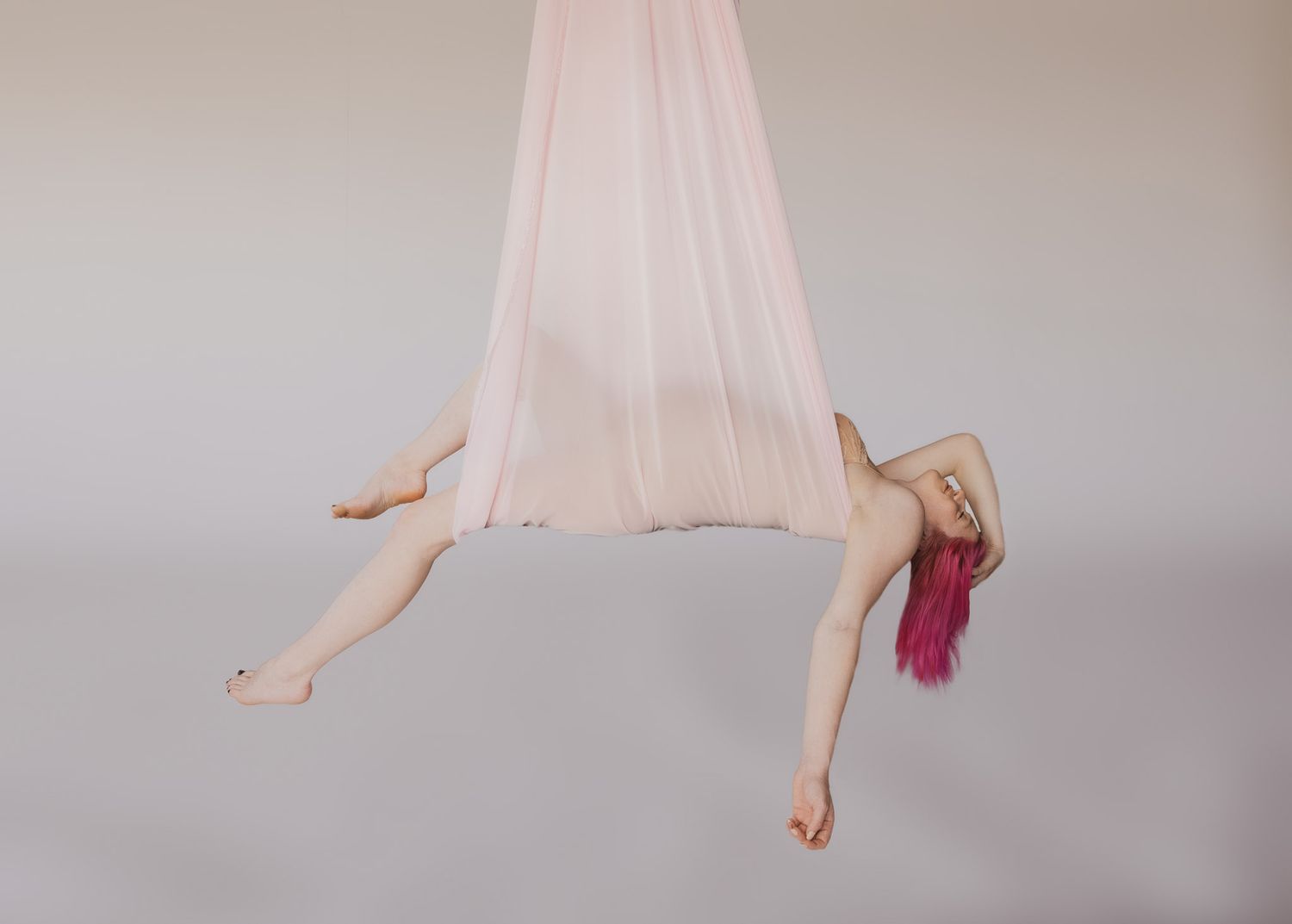 Aerial silk performer with pink hair gracefully poses suspended in pale pink fabric against gray backdrop.