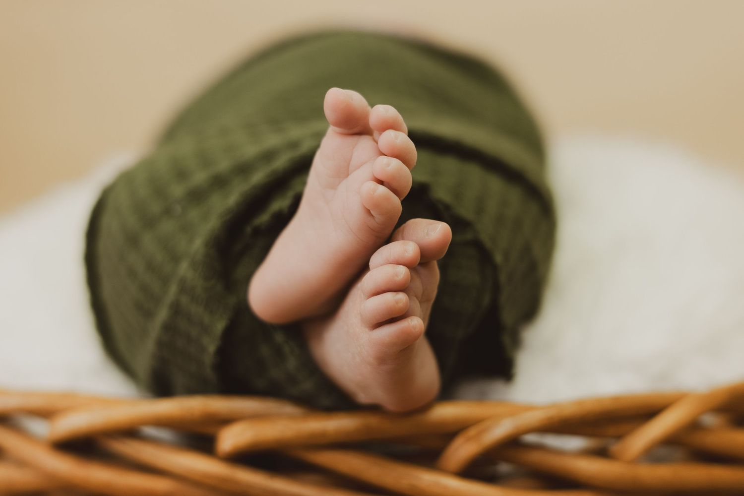 Tiny newborn baby feet peeking out from green swaddle blanket over edge of wicker basket.